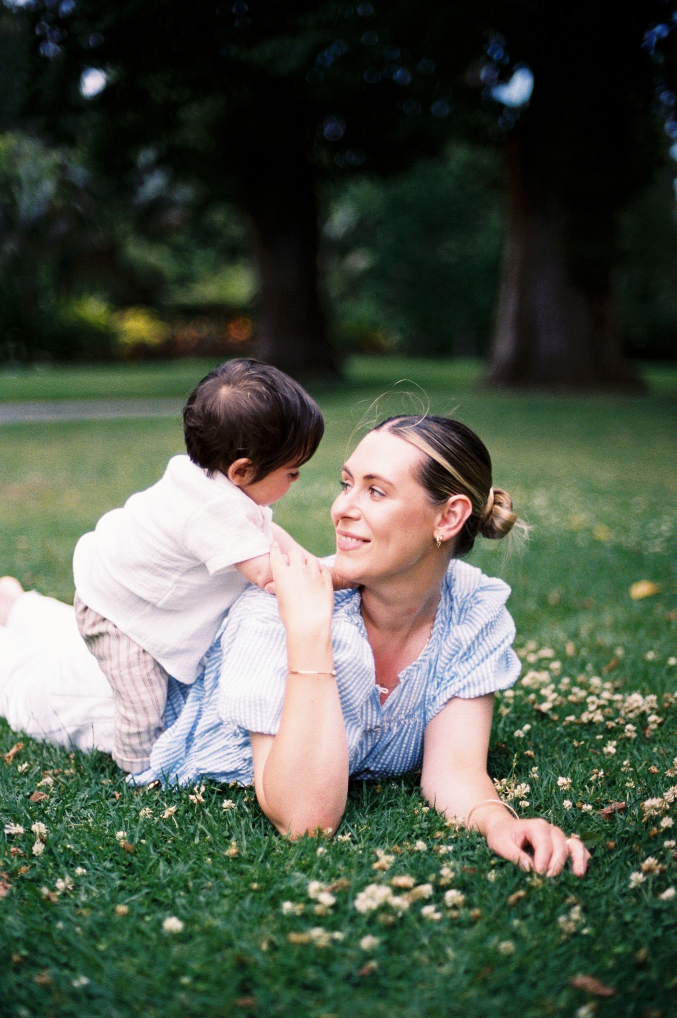 A parent and child lie together on grass with white dandelions while sharing a tender moment outdoors.