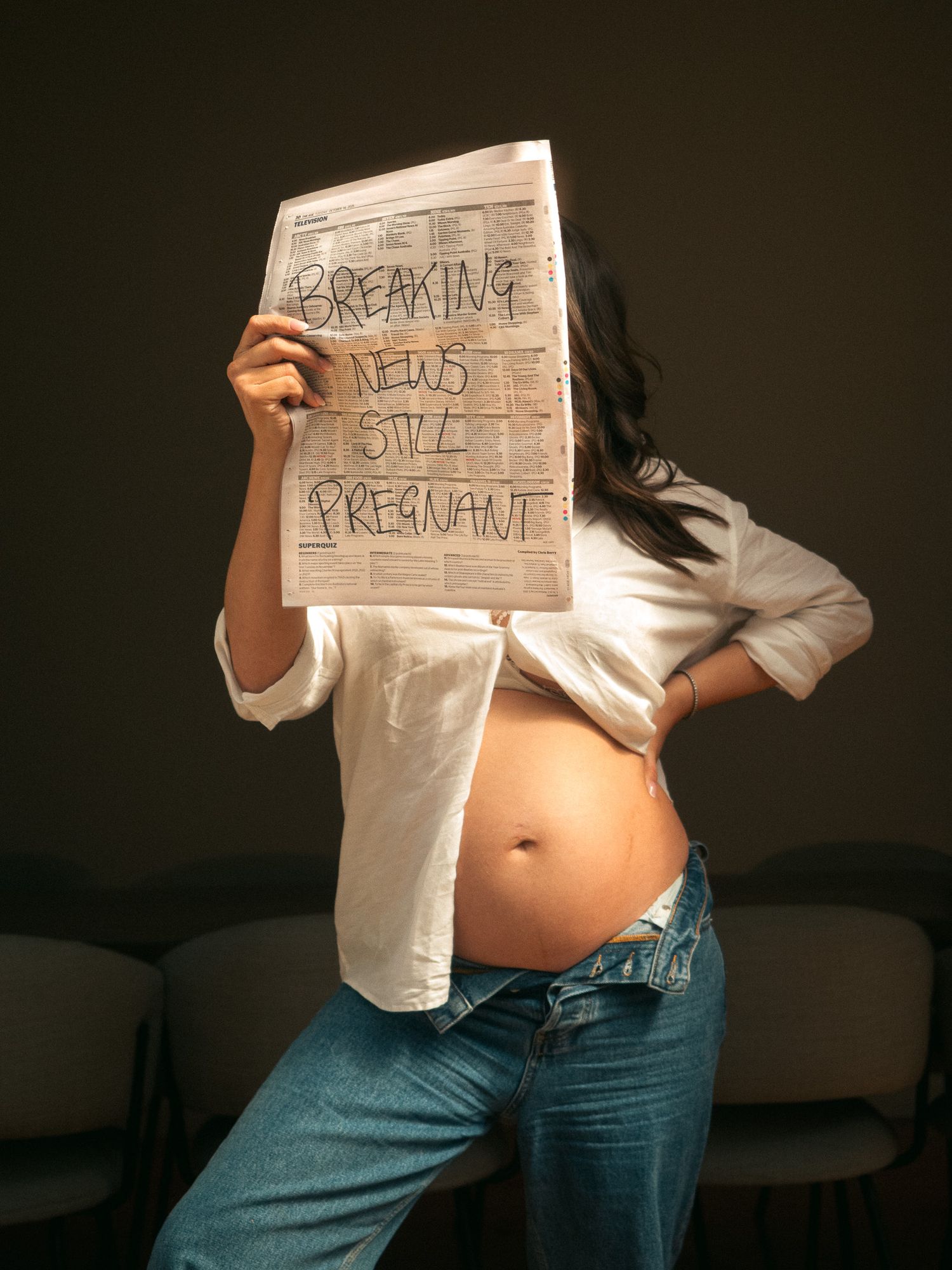 A creative maternity photoshoot with the pregnant woman standing and posing with a newspaper that says 'breaking news still pregnant'