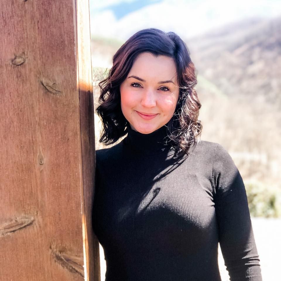 A person in a black turtleneck sweater poses outdoors against a wooden post with mountains in the background.