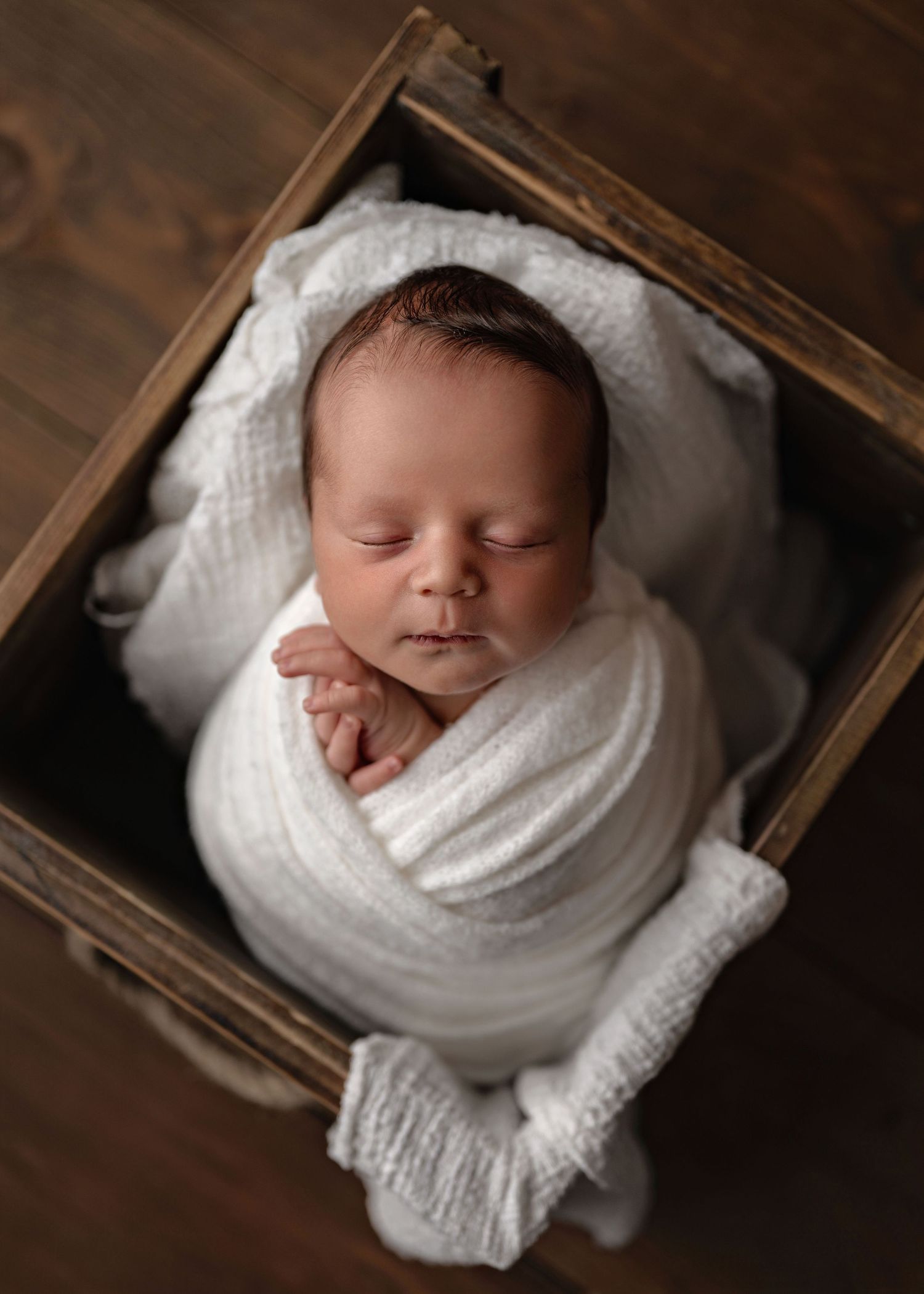 Sleeping newborn baby swaddled in white wrap, nestled in a rustic wooden crate on a dark wood floor.