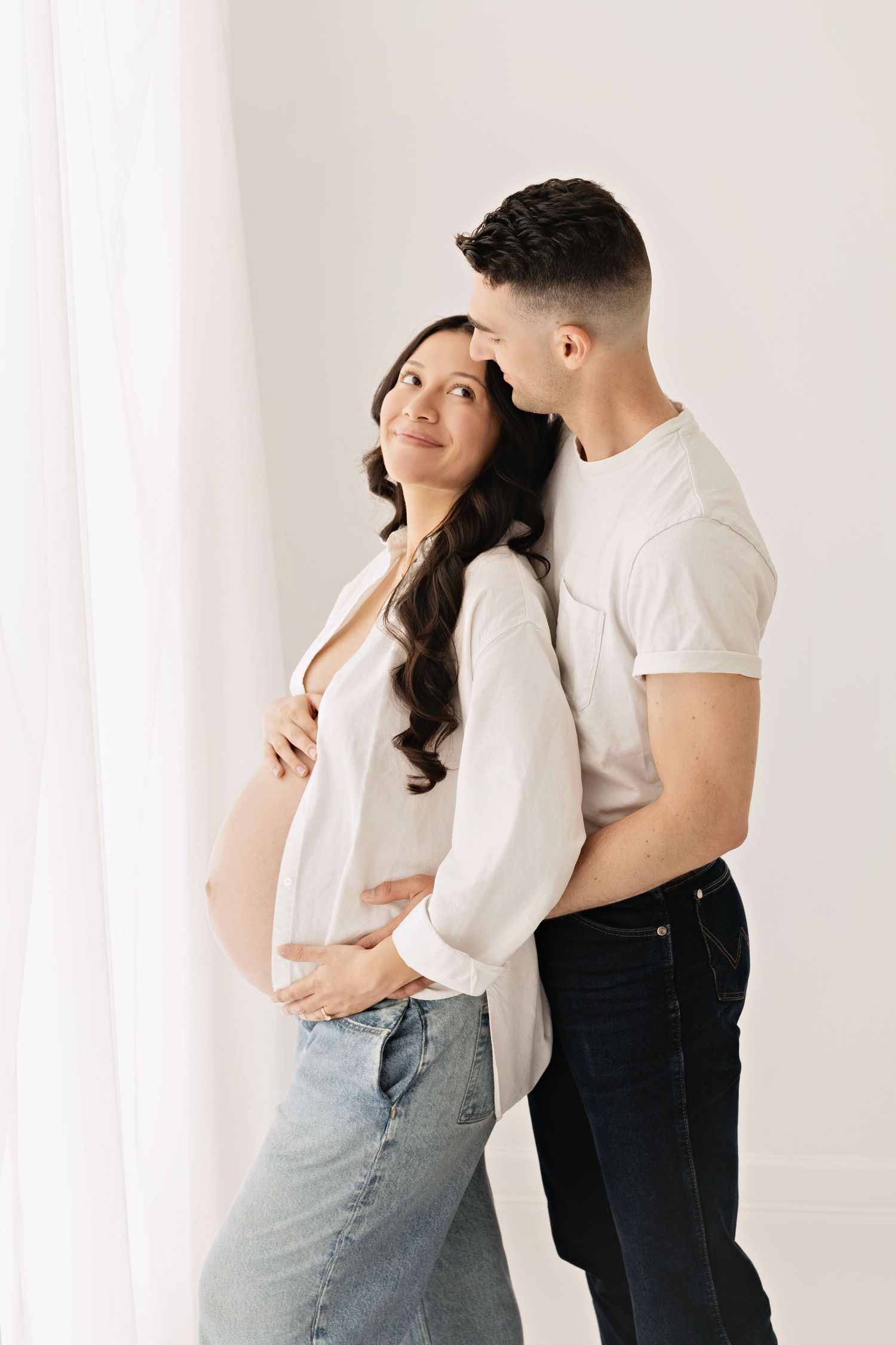 A man tenderly embraces a pregnant woman in white shirt and jeans, standing together by a bright white curtain.