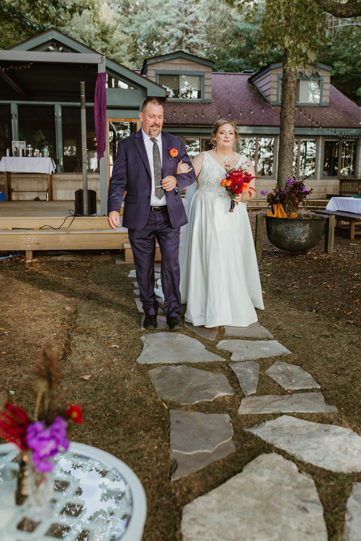 Wedding couple stands on stone pathway surrounded by trees with red floral accents in outdoor venue setting.