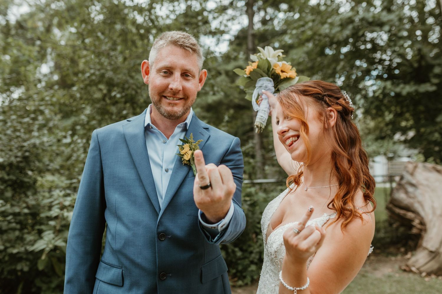 Smiling bride and groom in blue suit showing off wedding rings outdoors with greenery background.
