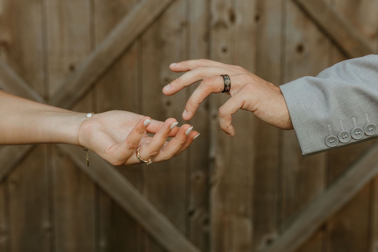 Two hands reaching toward each other, one wearing a ring, against a rustic wooden barn door background.