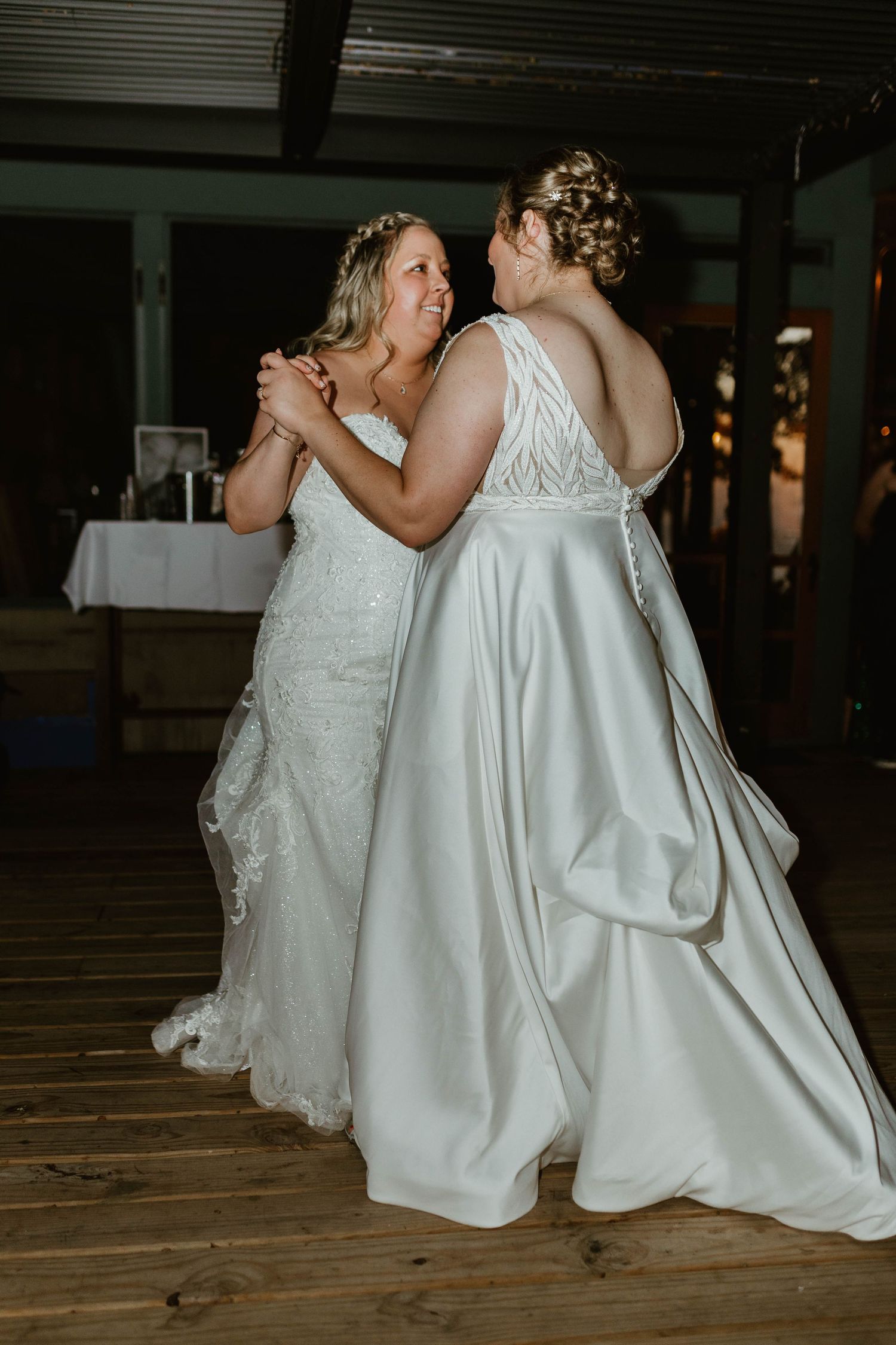 Two people in white wedding dresses sit together on rustic wooden floor during reception.