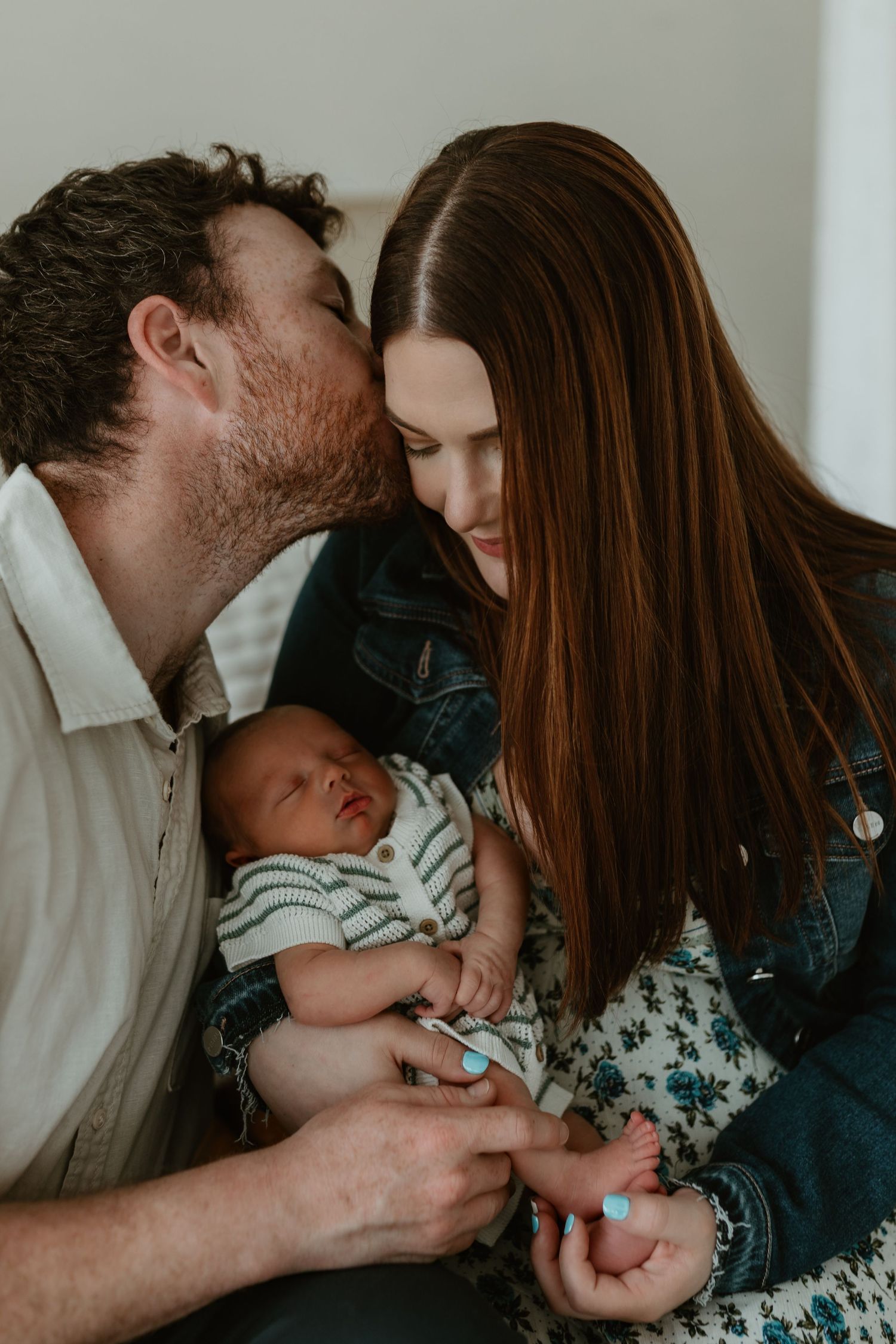 New parents sharing a tender moment, father kissing mother's cheek as they hold their sleeping newborn baby together.