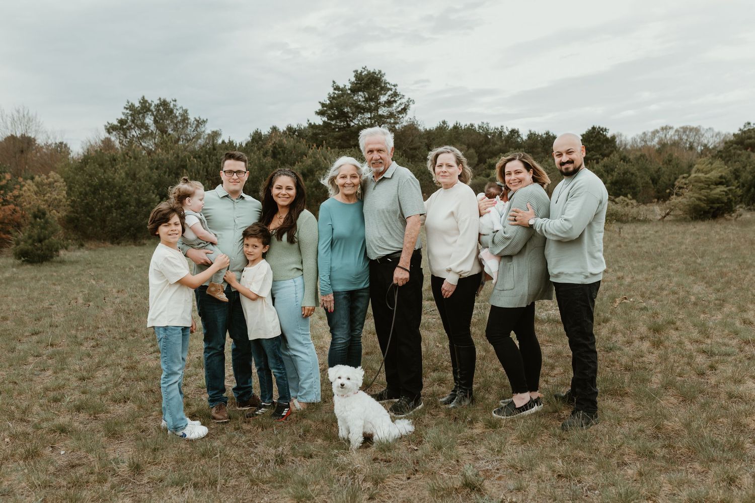 Multi-generational family portrait outdoors in a field, wearing coordinated mint and neutral tones, with a small white dog.