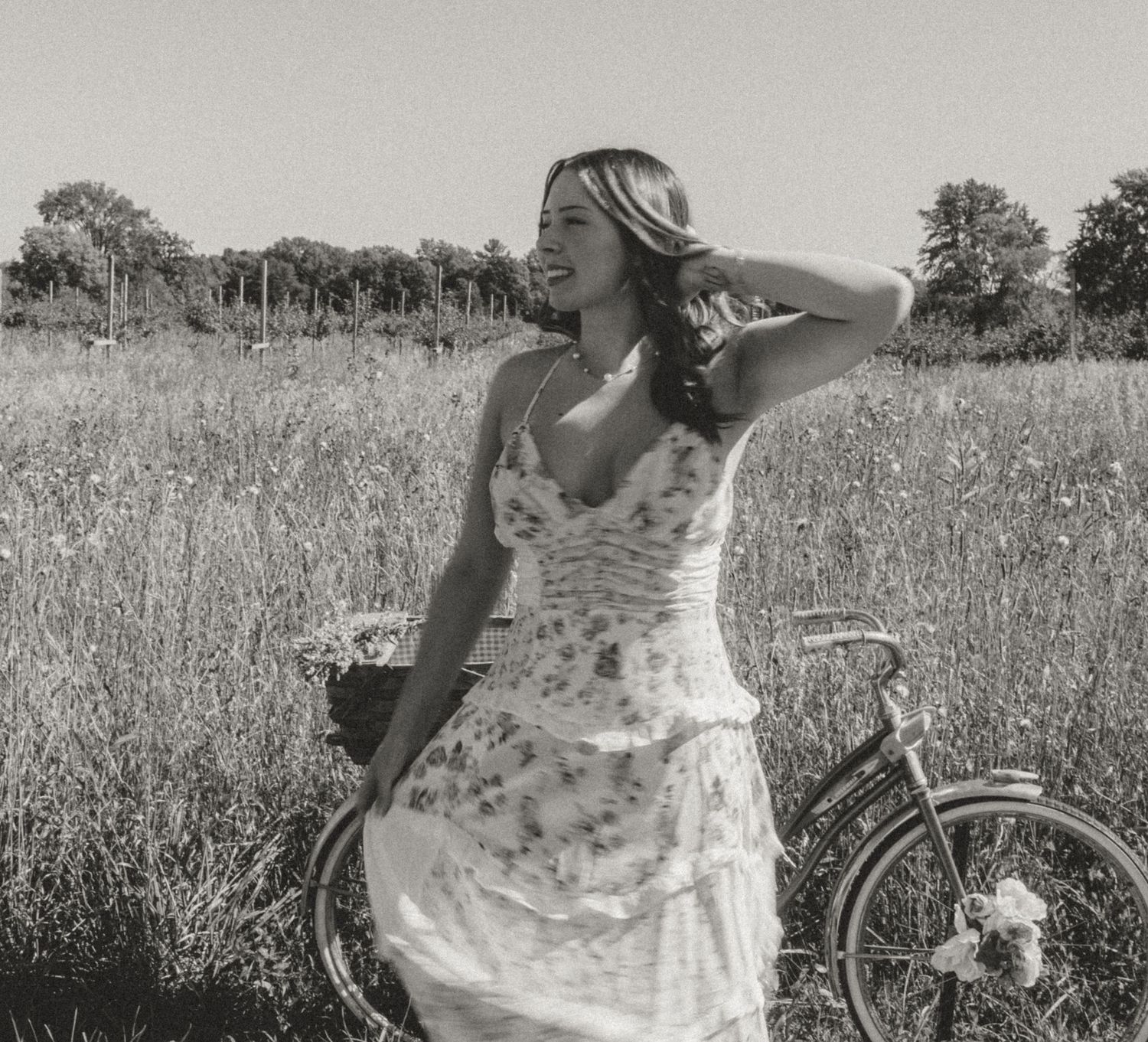 Black and white photo of a smiling woman in a floral dress standing in a meadow beside a vintage bicycle.