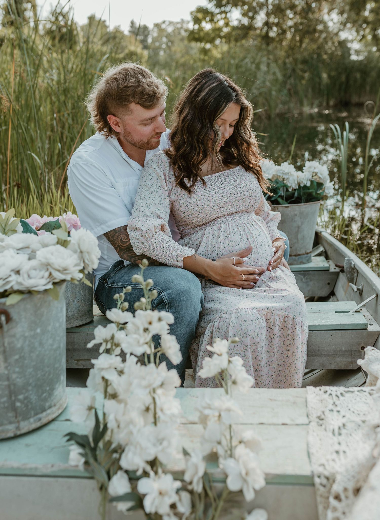 Couple sharing a tender maternity photo shoot moment surrounded by white flowers near a serene pond.