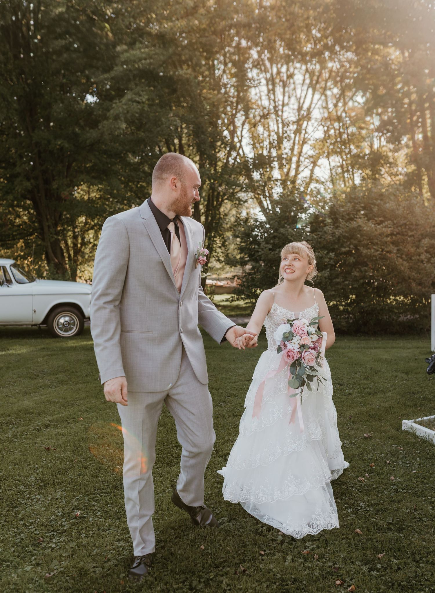A couple in wedding attire walks through a sunlit garden during golden hour.