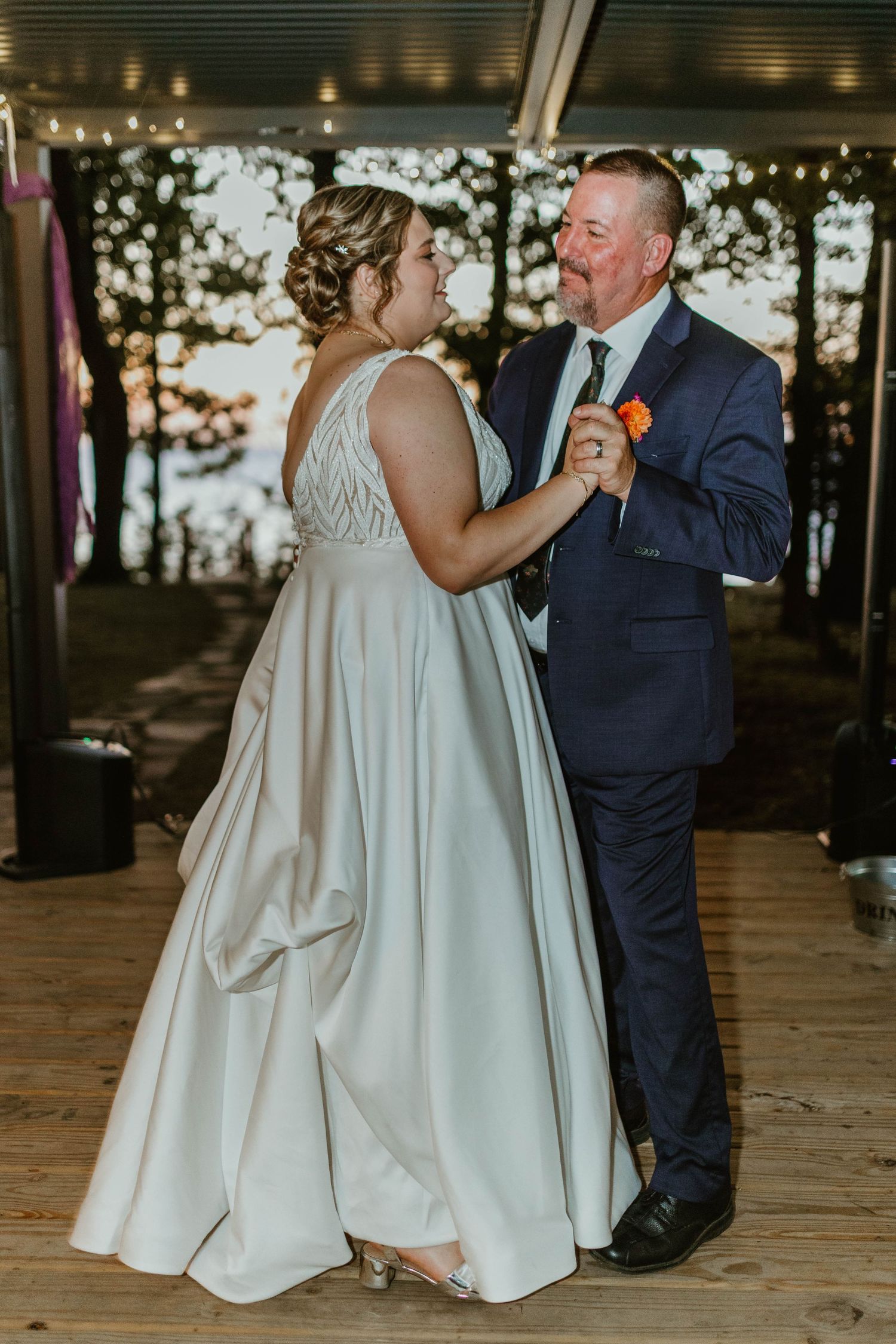 Wedding couple shares intimate dance moment on wooden deck with forest backdrop at sunset.