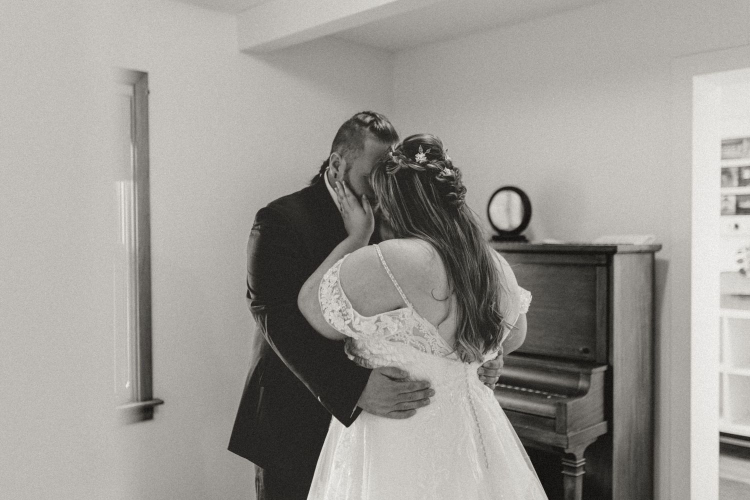 Black and white photo of a bride and groom sharing a kiss near a vintage piano on their wedding day.