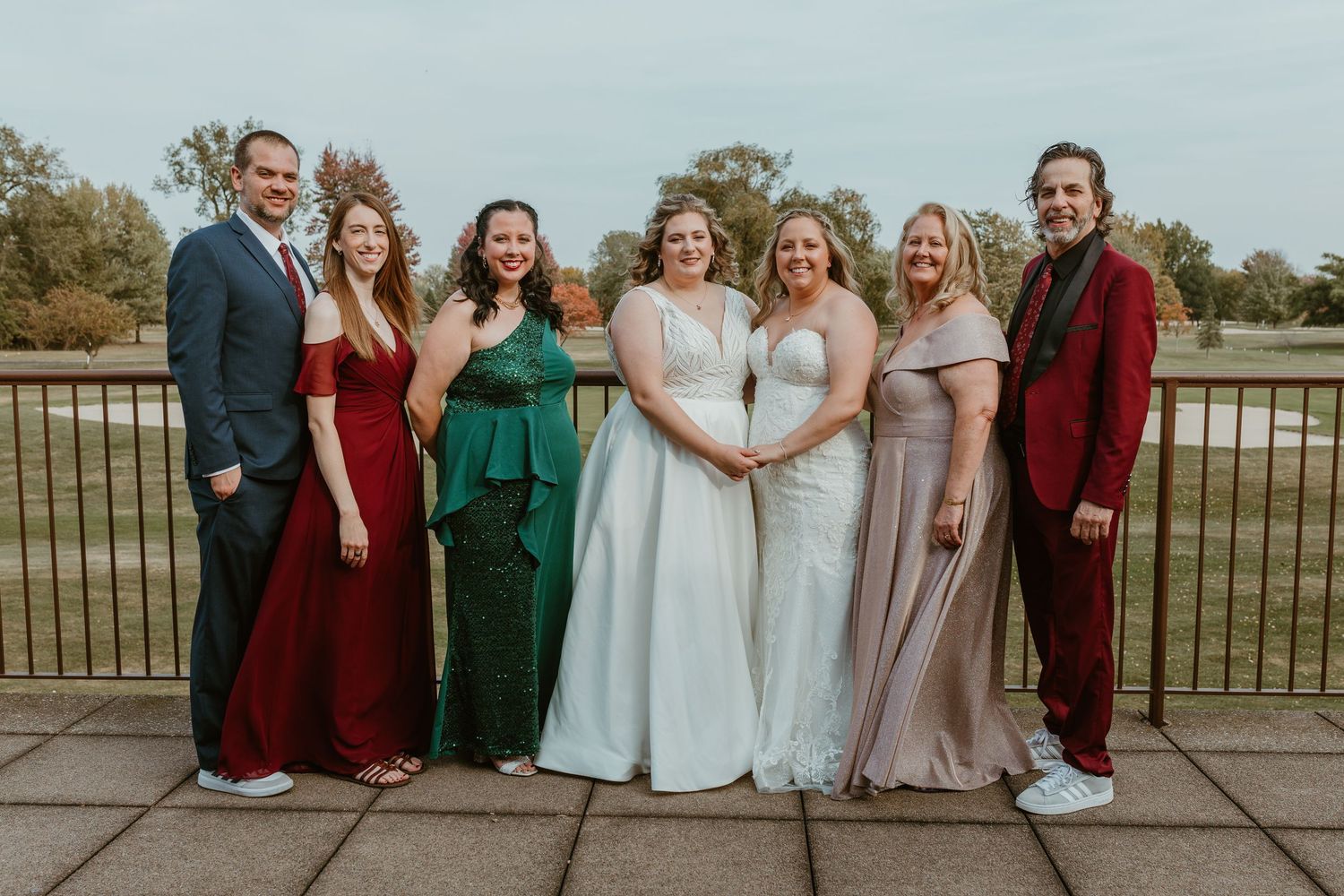 Wedding party poses together on outdoor deck wearing burgundy, green, and neutral colored attire.