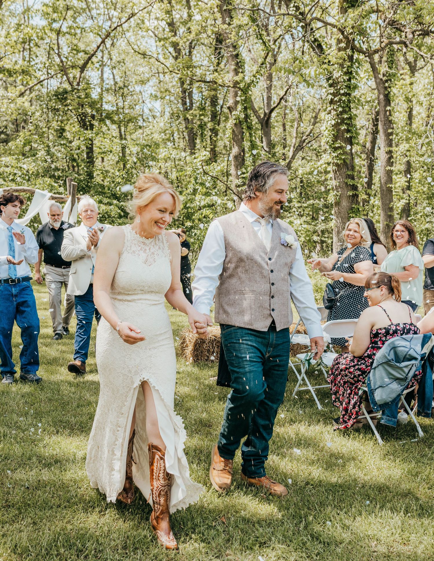 A couple walking hand in hand at their outdoor woodland wedding with guests in background.