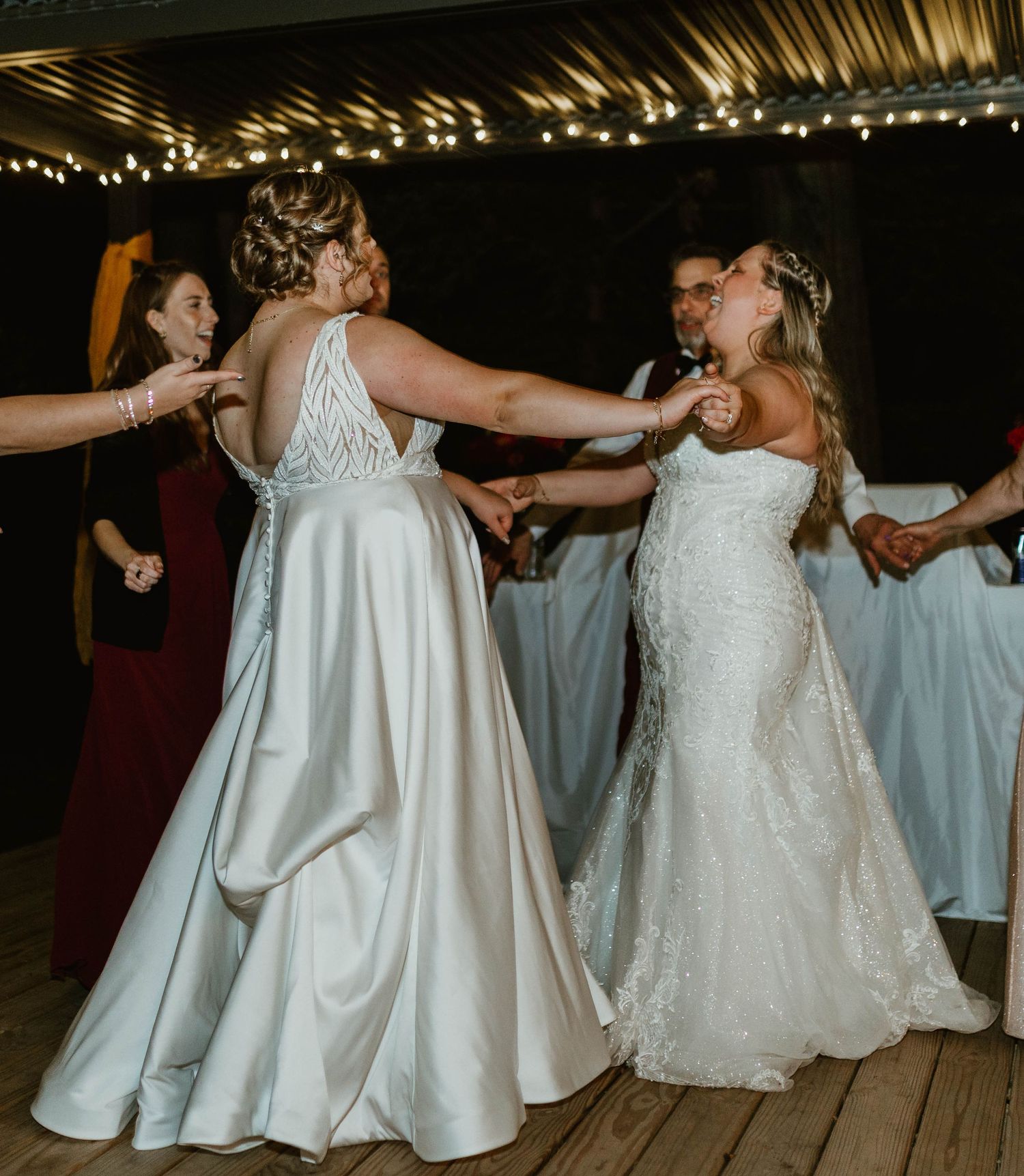 Wedding party dances in circle on wooden dance floor wearing mix of white and colorful dresses.