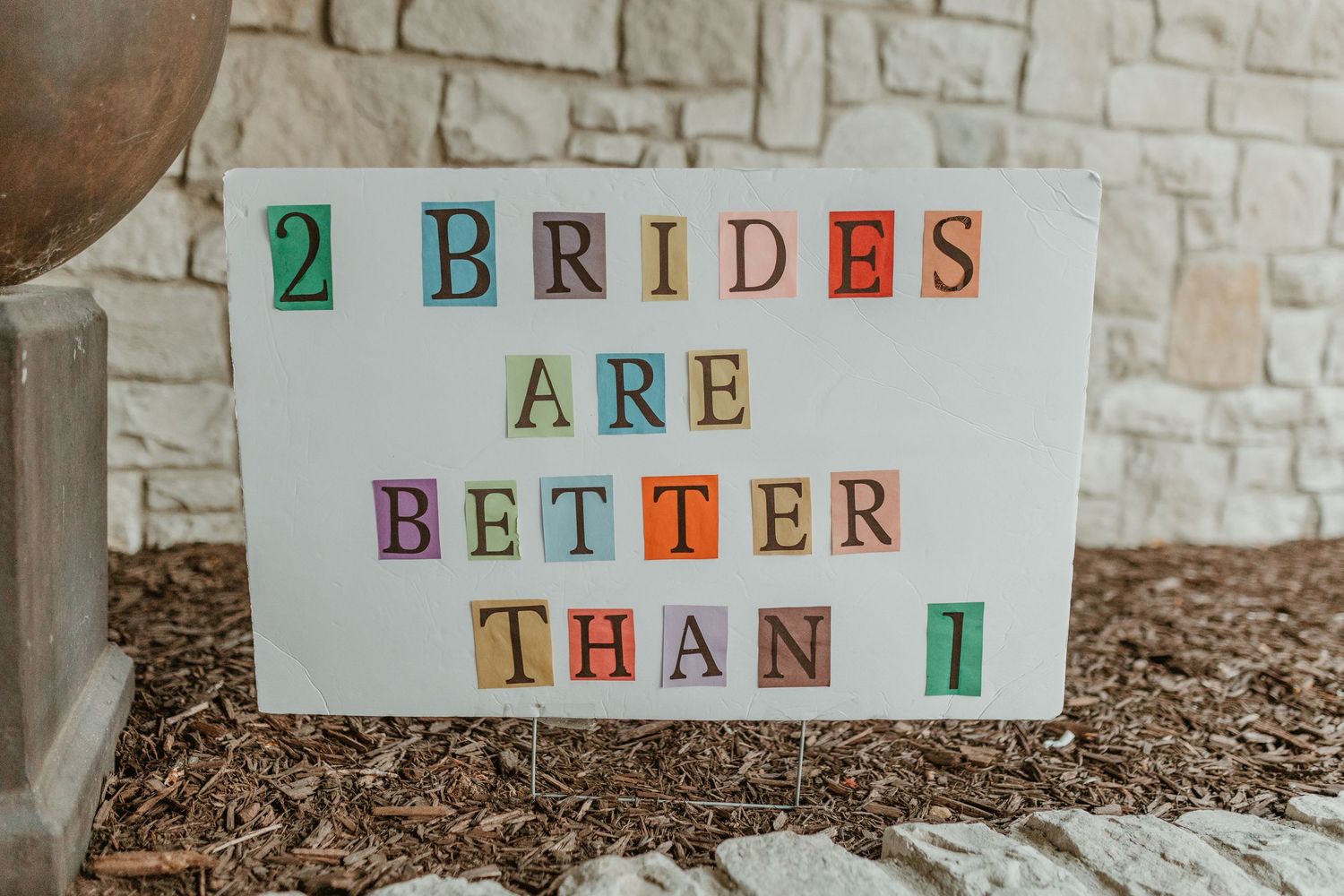 A decorative sign reading '2 Brides are Better than 1' displayed against a stone wall.