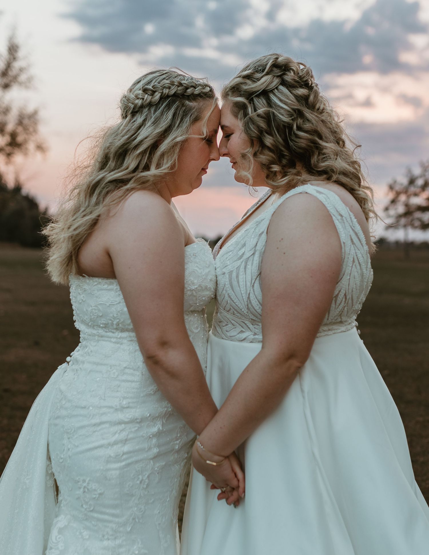 Two brides in white wedding dresses share an intimate moment at sunset in a field.