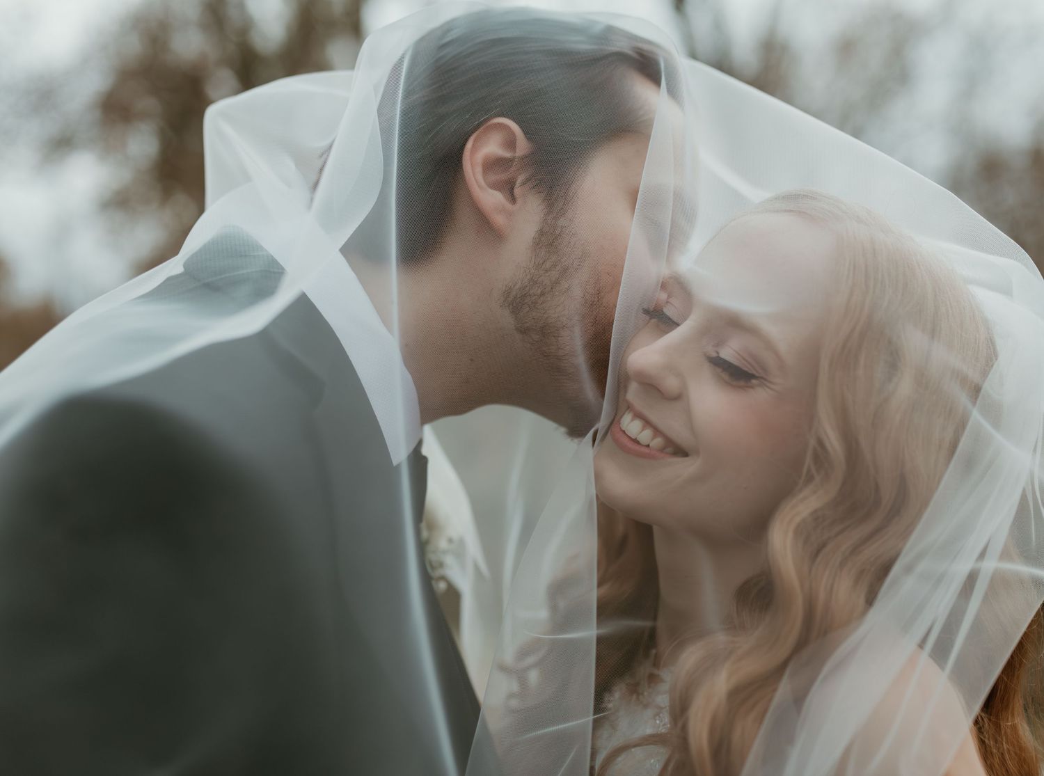 A bride and groom share a romantic moment under a flowing white wedding veil outdoors.