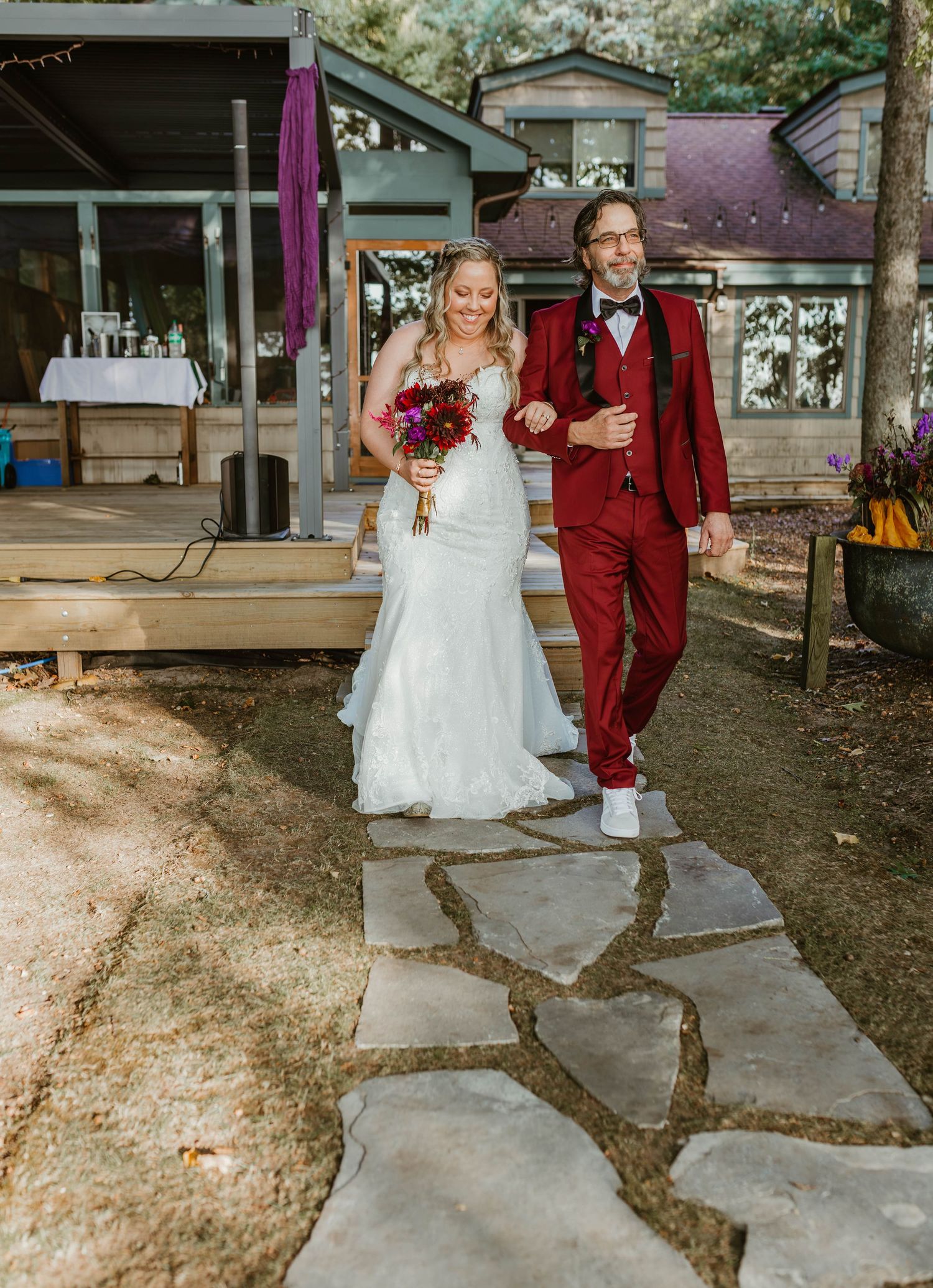 Wedding couple walks along stone pathway with one wearing a red suit and the other in white dress.