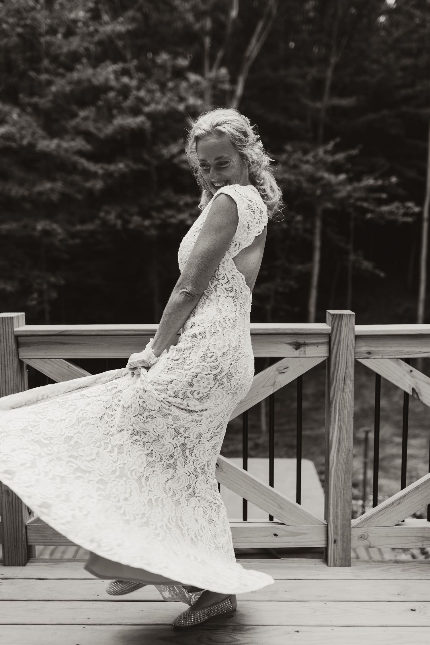 A flowing white lace wedding dress twirls gracefully on a wooden deck in black and white.