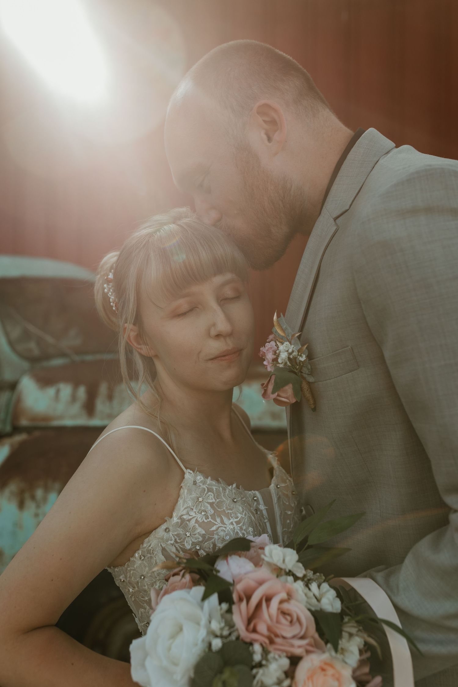 A bride and groom share a tender moment bathed in golden sunlight, she holds a pink and white floral bouquet.