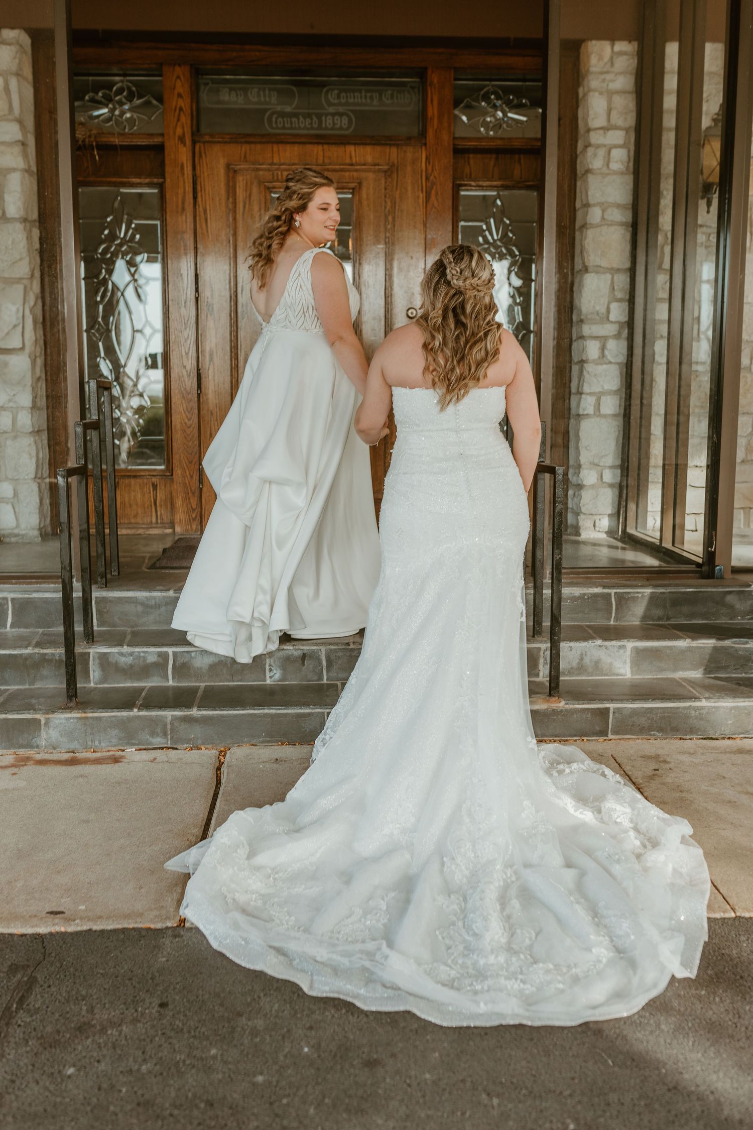 Two people in elegant white wedding dresses stand on stone steps in front of wooden church doors.
