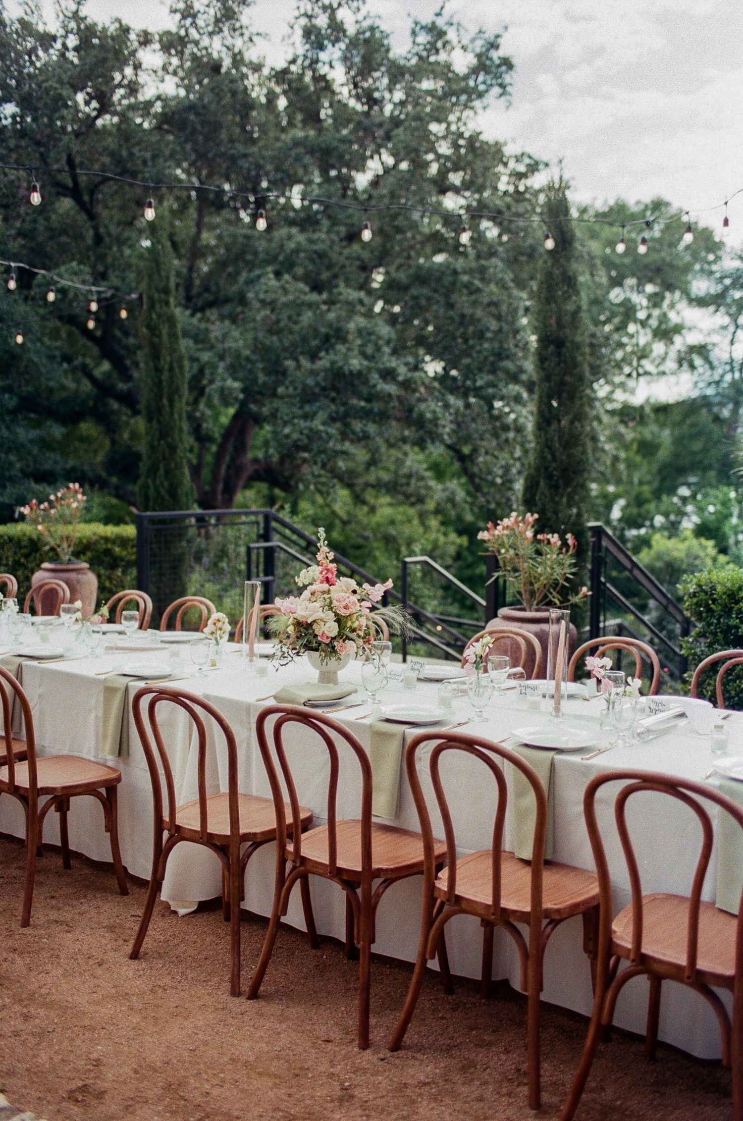 Elegant outdoor wedding reception table with white linens and wooden bentwood chairs set beneath string lights and cypress trees.