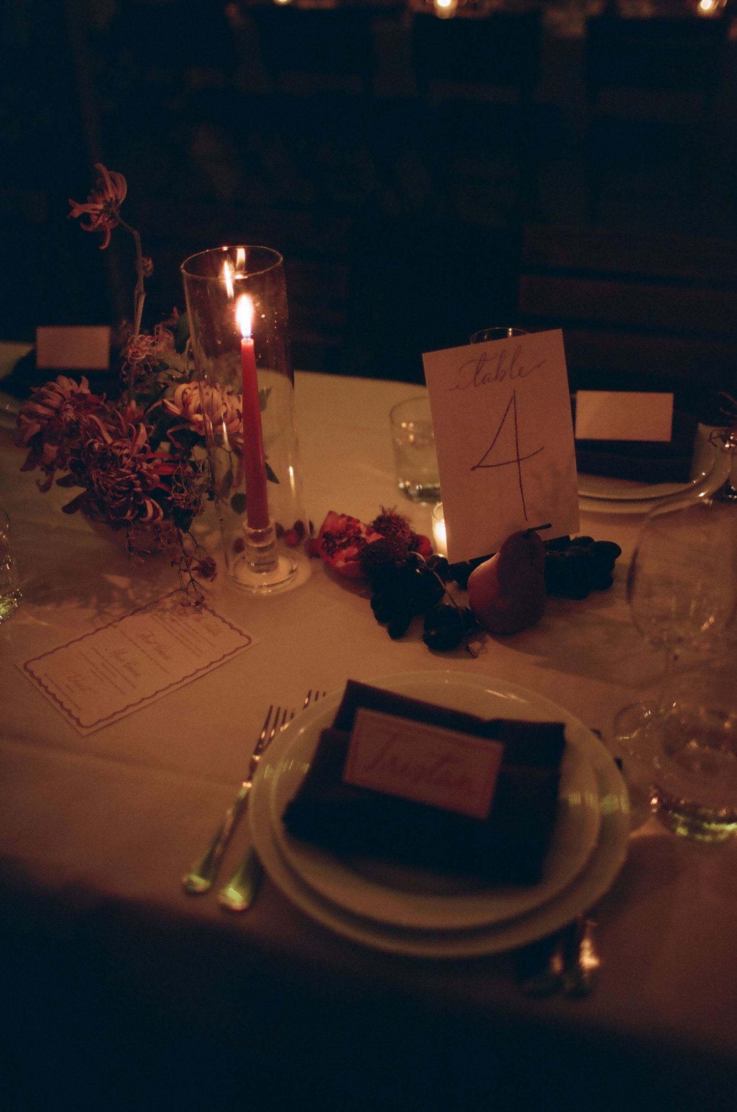 Candlelit table settings with white plates and floral centerpieces create an elegant dining atmosphere in dim lighting.