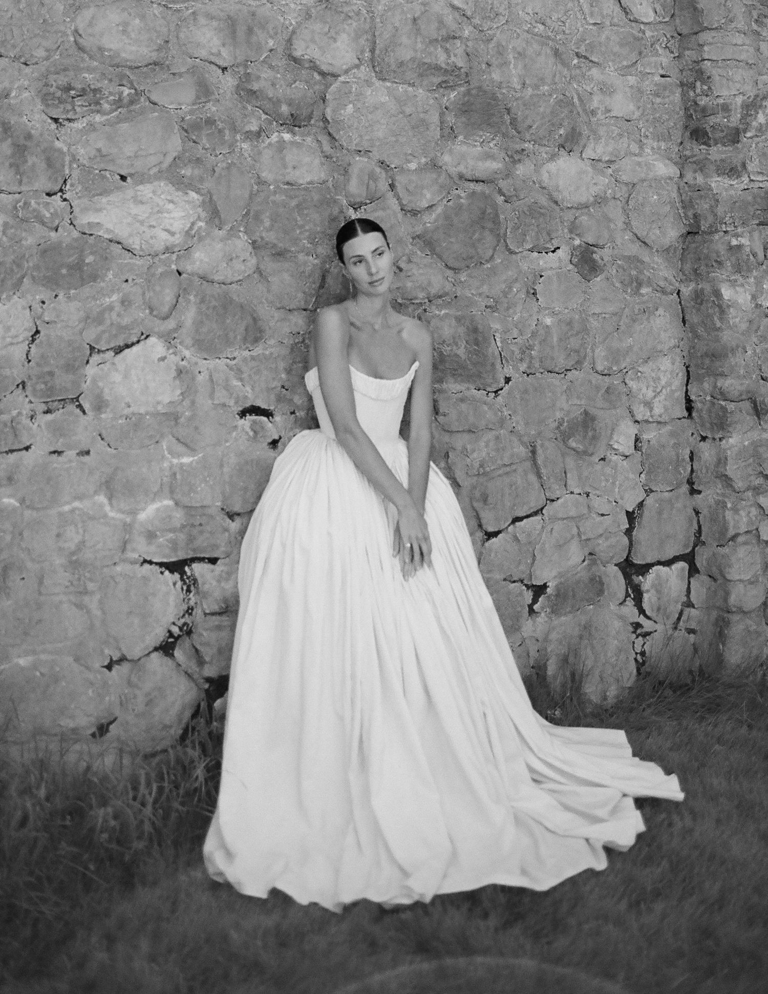Black and white photo of a bride in a strapless ball gown wedding dress posed against a rustic stone wall.