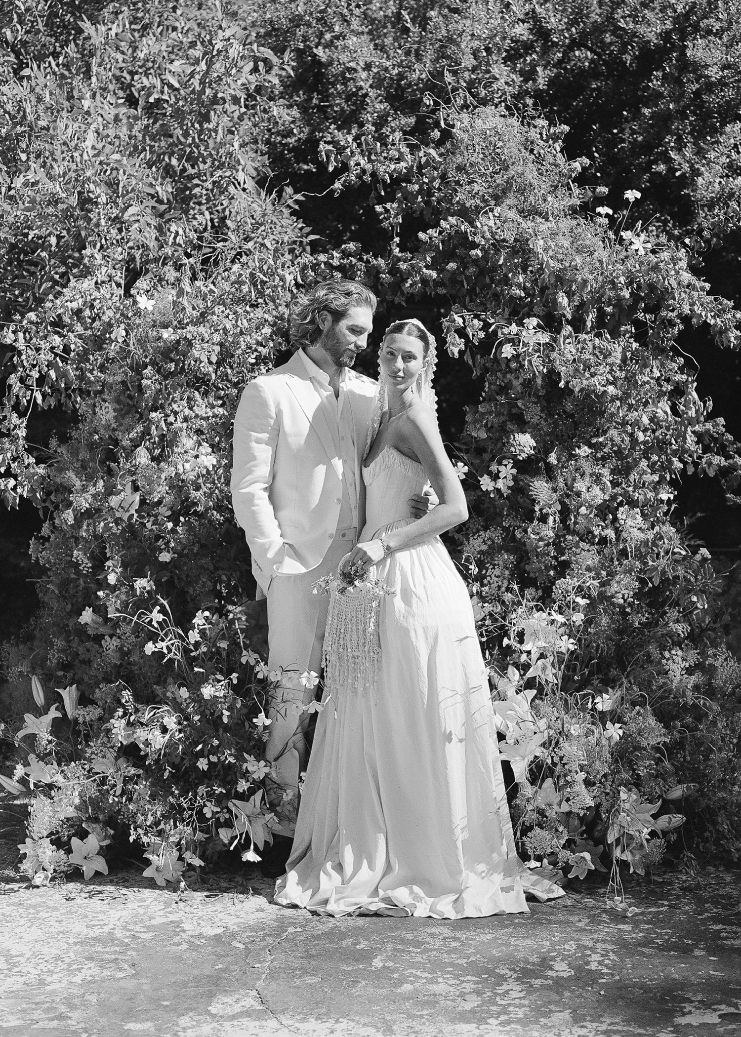 Black and white wedding photo of a couple in formal attire standing before lush greenery.
