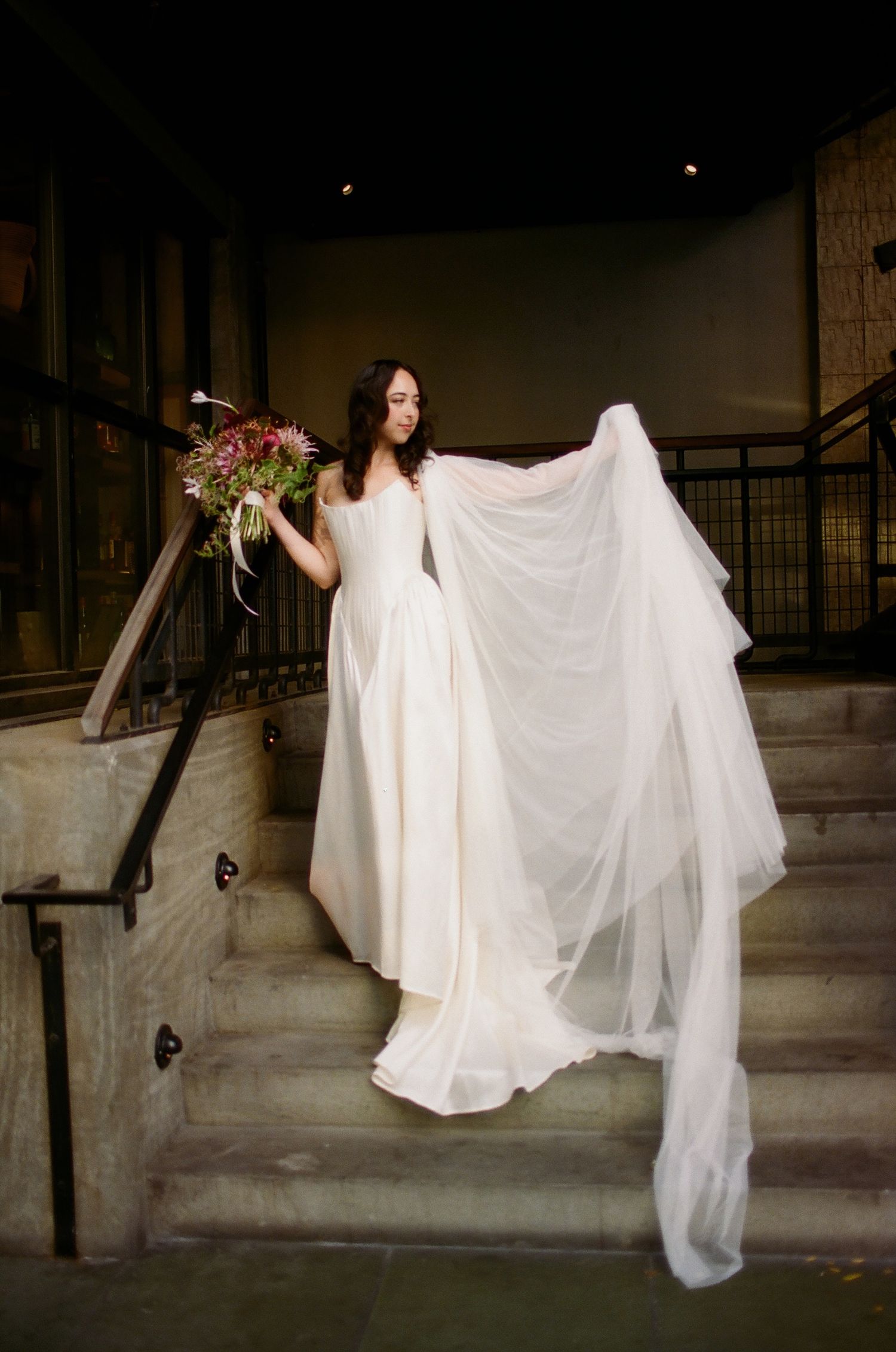 Bride in flowing white gown with veil holding floral bouquet on stone steps in elegant venue.