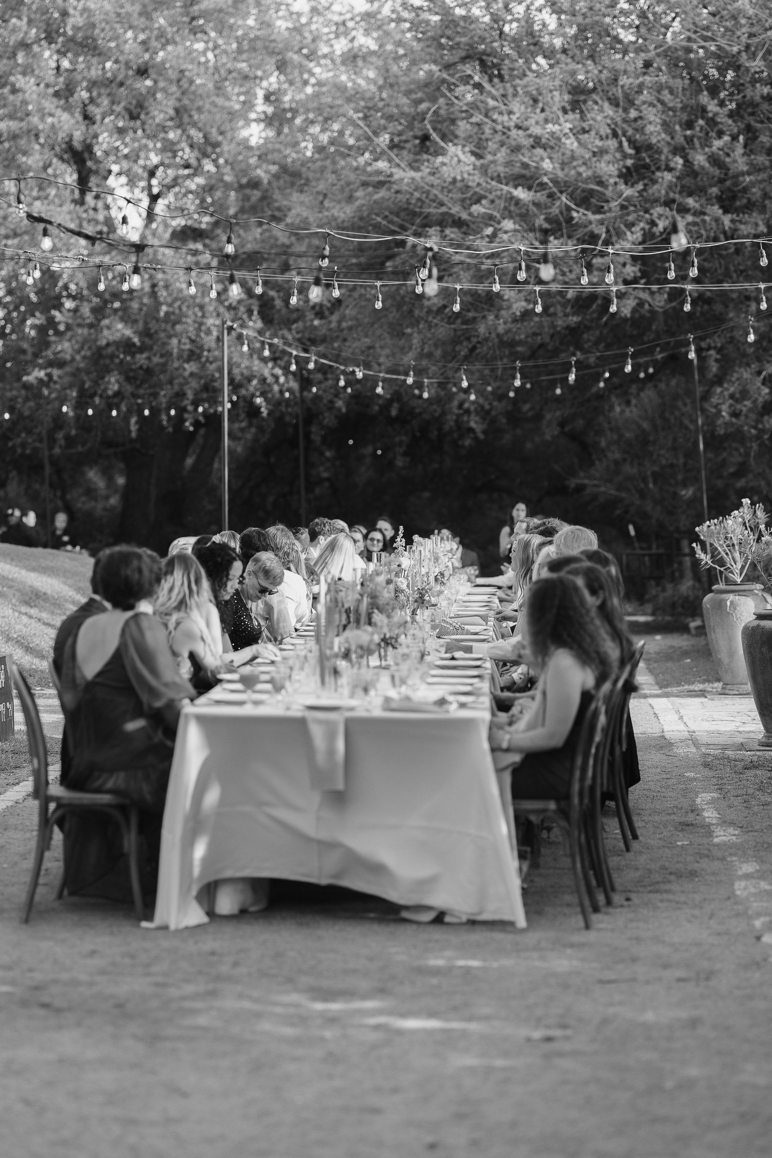 Black and white outdoor wedding reception with guests seated at long decorated table under string lights amid trees.