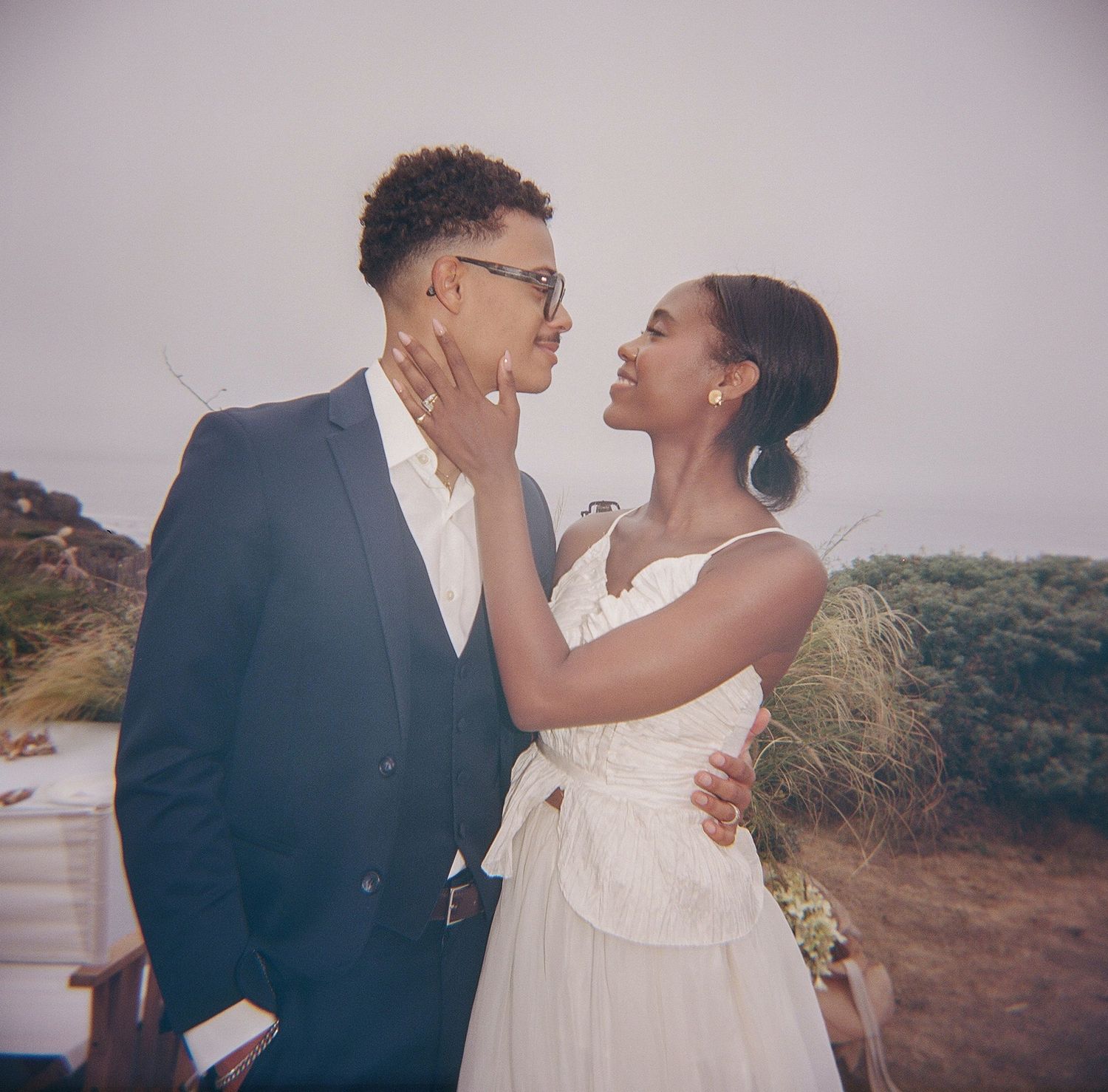 A couple in wedding attire shares a romantic moment outdoors, the bride in white and groom in a navy suit.
