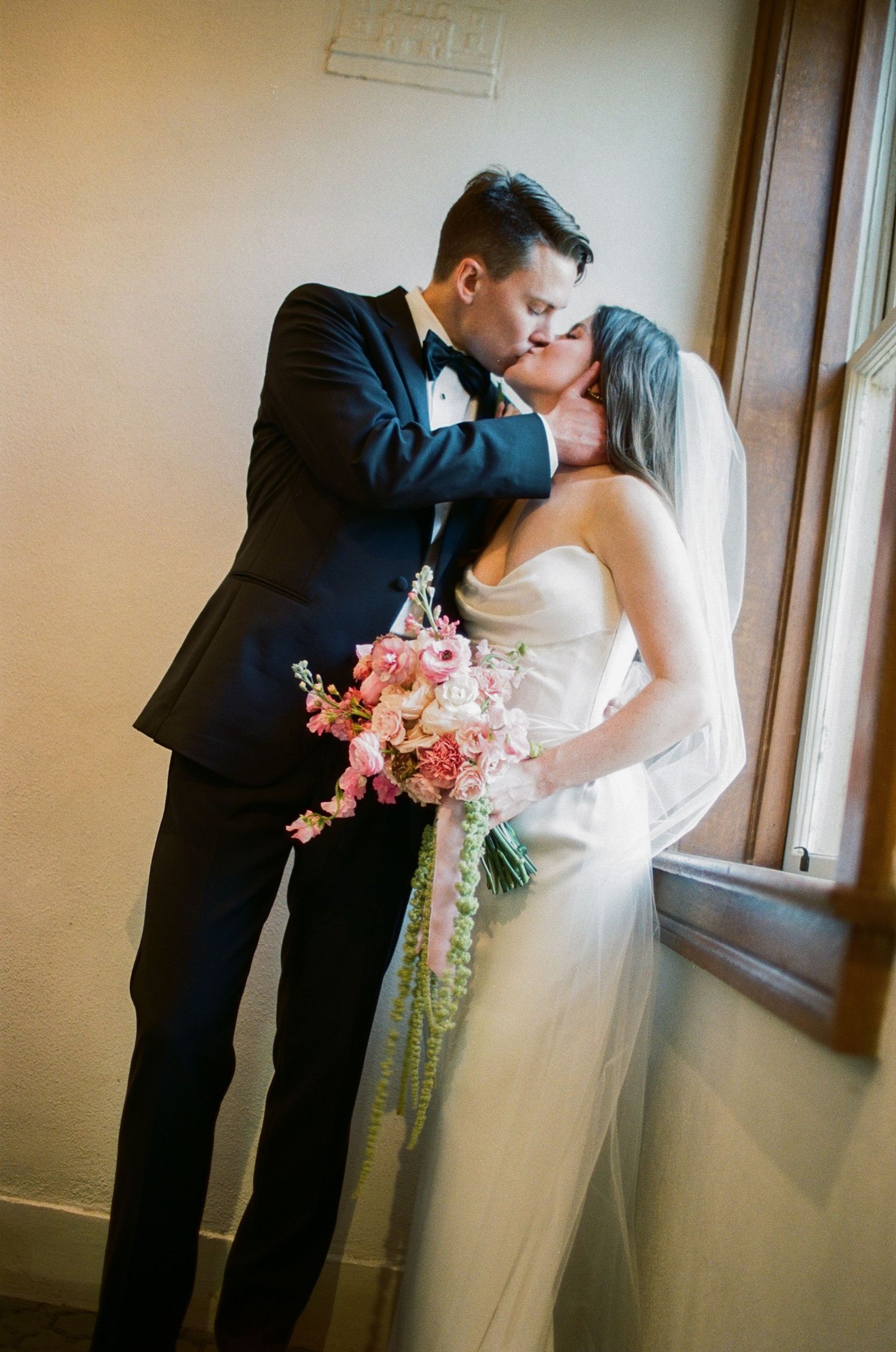 A romantic wedding moment captured near a window with soft natural lighting.