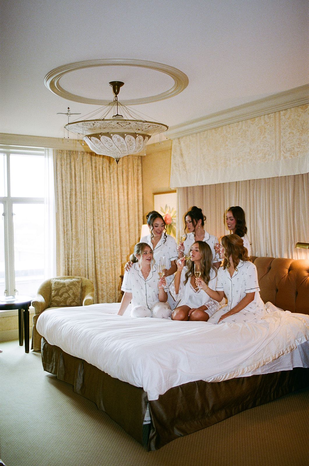 Group wearing white robes relaxing together on large hotel beds in an elegant room with chandelier.