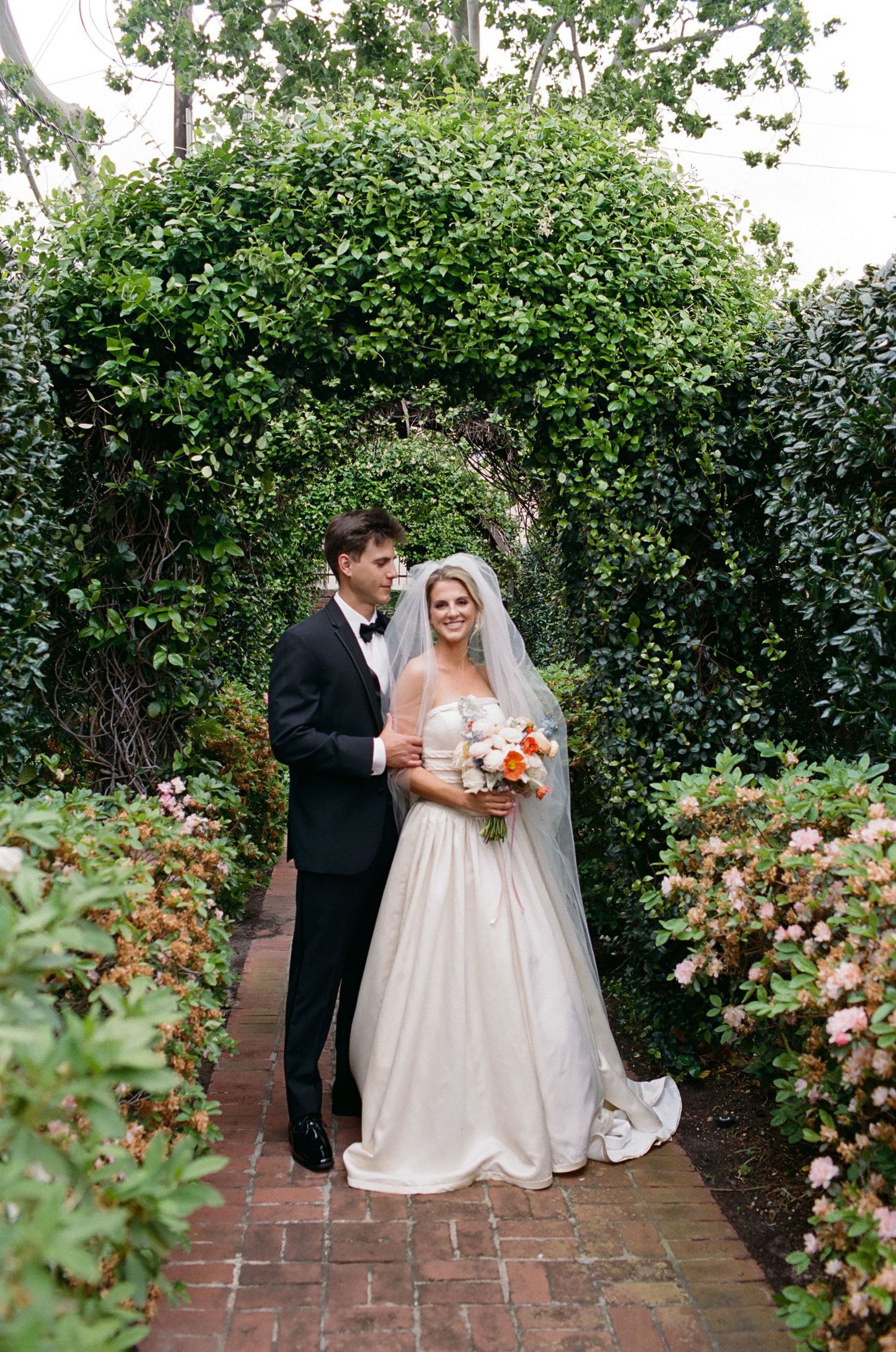 Wedding couple poses in a lush garden pathway with blooming flowers.