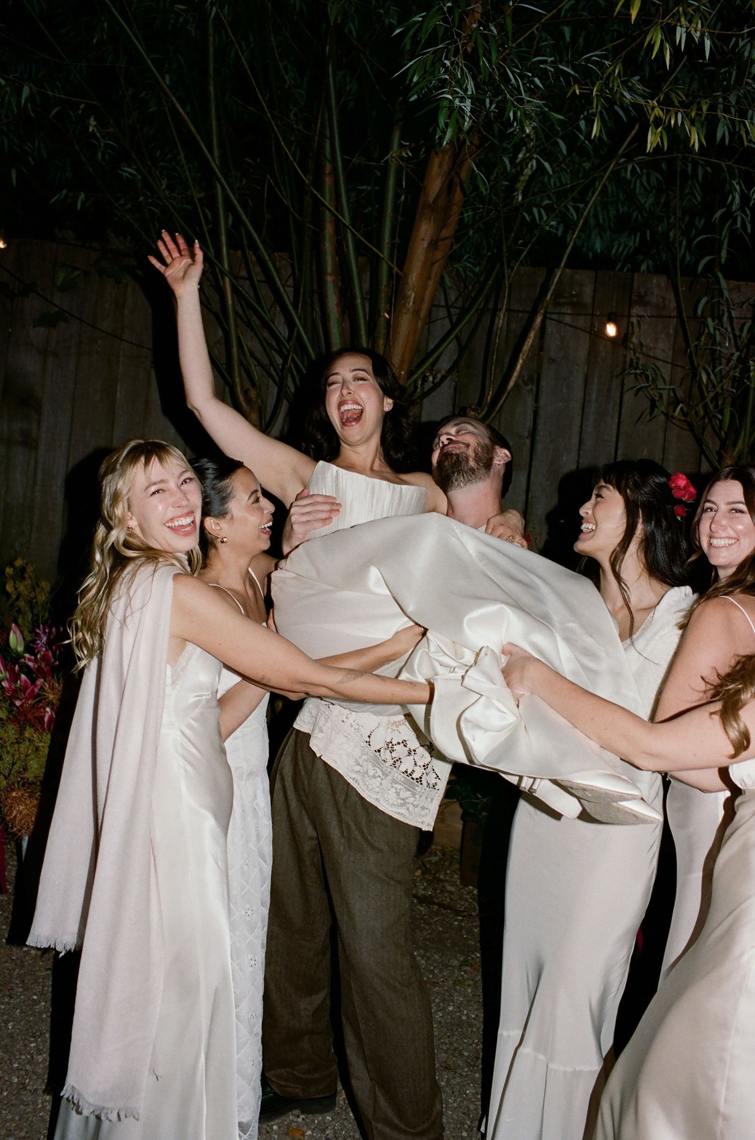 Wedding party in white attire dancing and celebrating at an outdoor evening reception with bamboo trees in background.