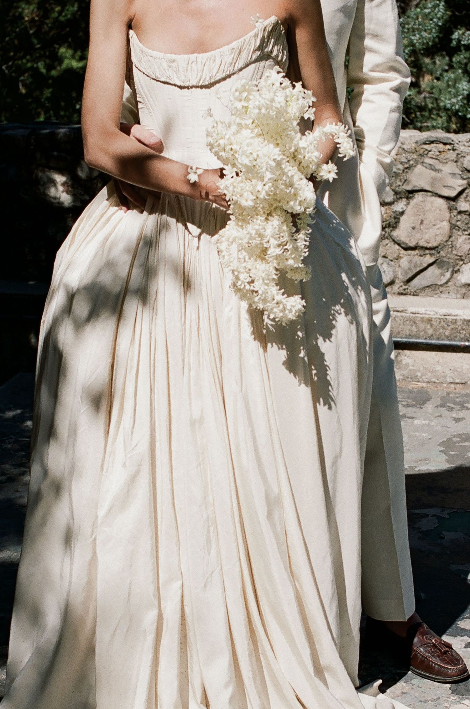 Bride in ivory strapless wedding gown holding white floral bouquet beside groom in cream suit outdoors.