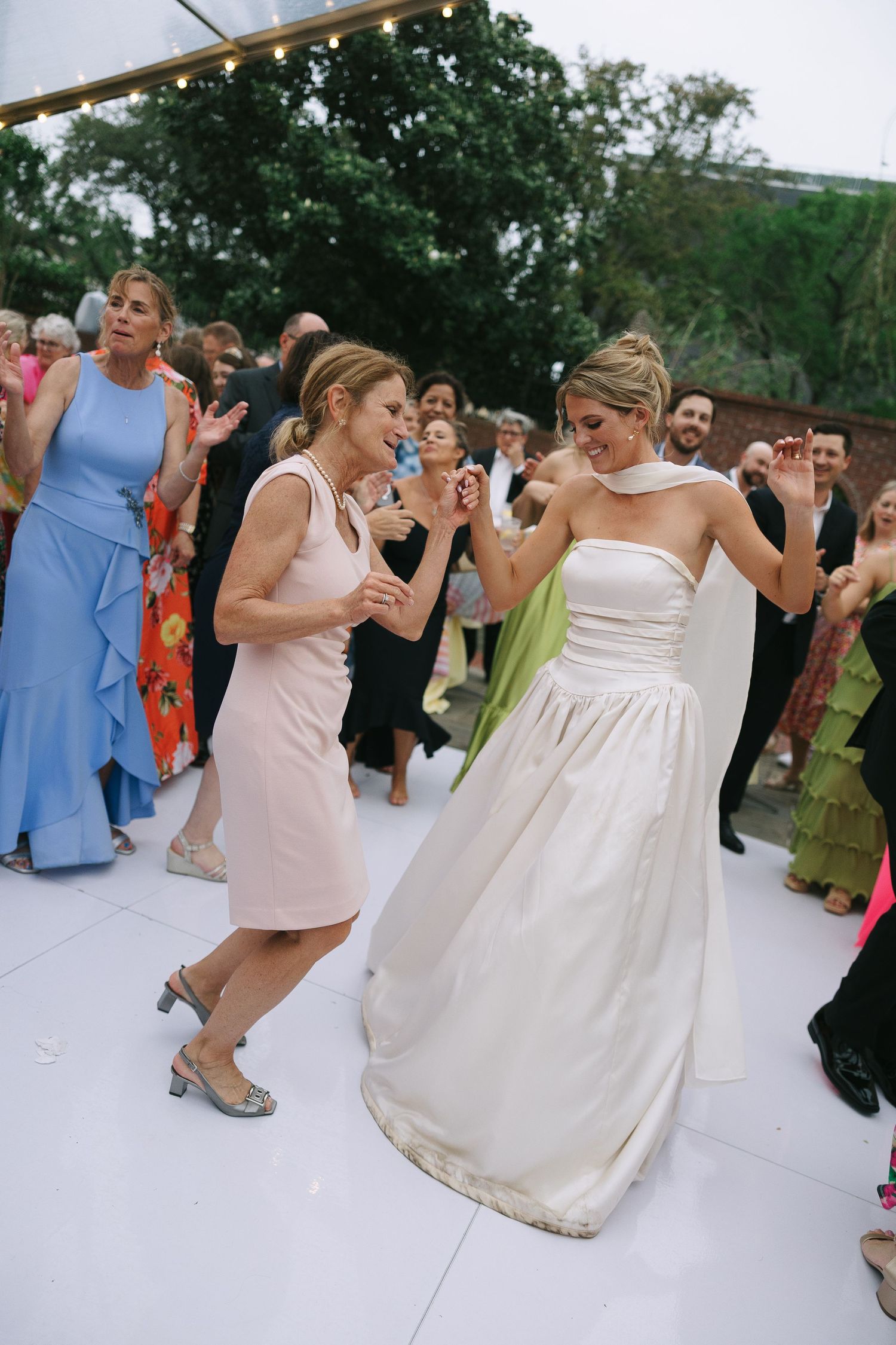 Wedding guests in colorful dresses dancing energetically on a white outdoor dance floor.