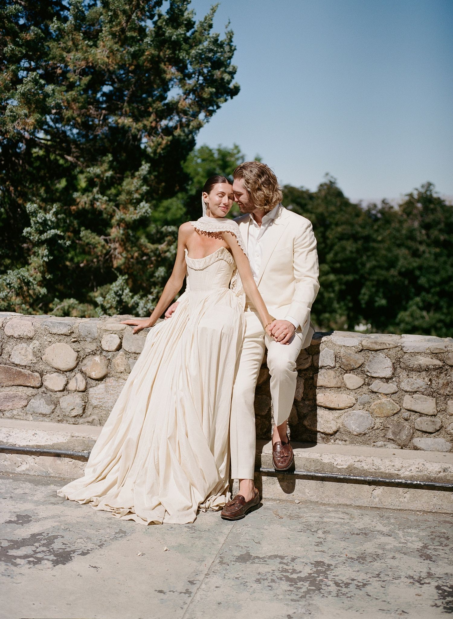 Couple in elegant cream wedding attire embracing on stone terrace surrounded by lush green trees on sunny day.