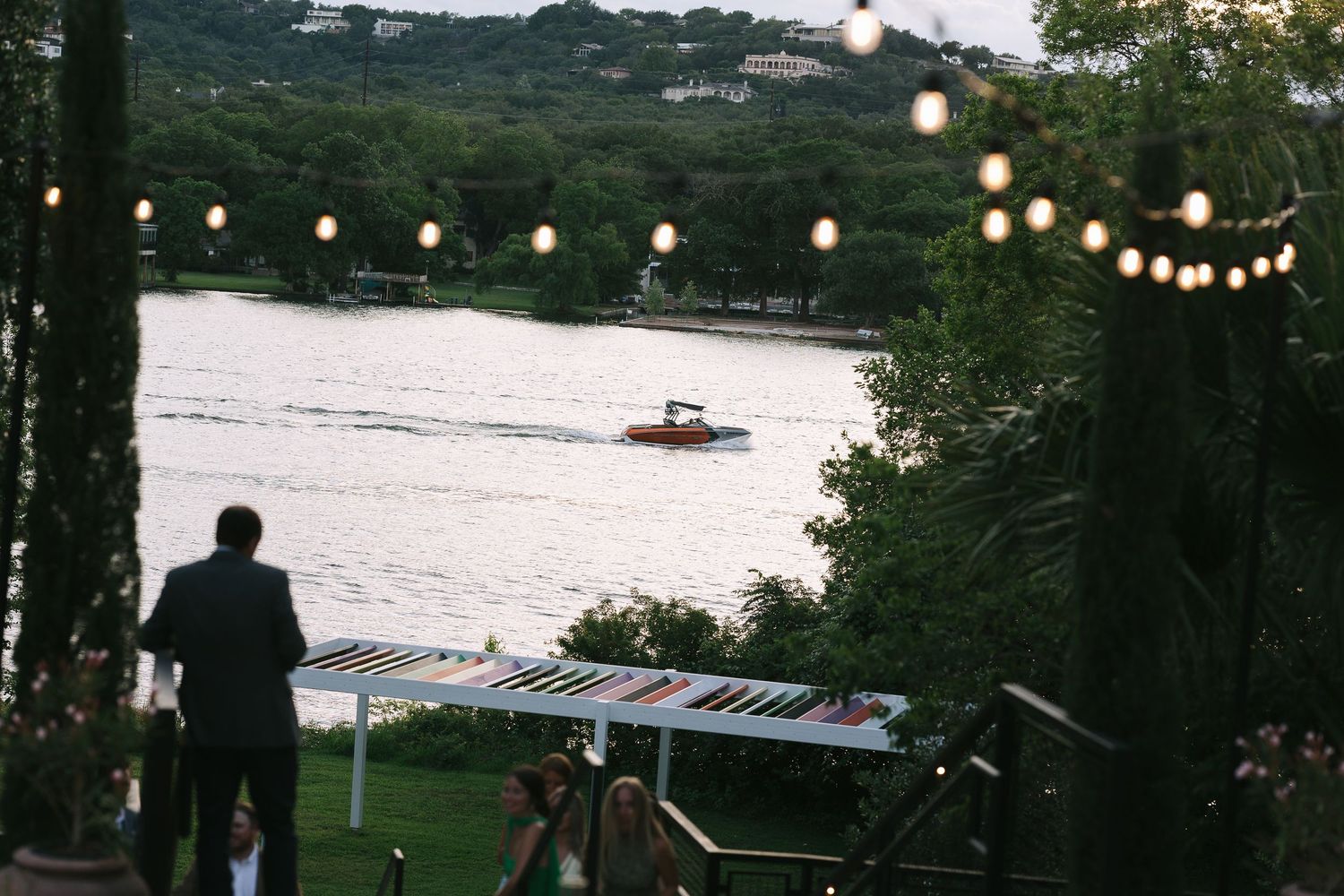 String lights illuminate an outdoor lakeside venue at dusk.