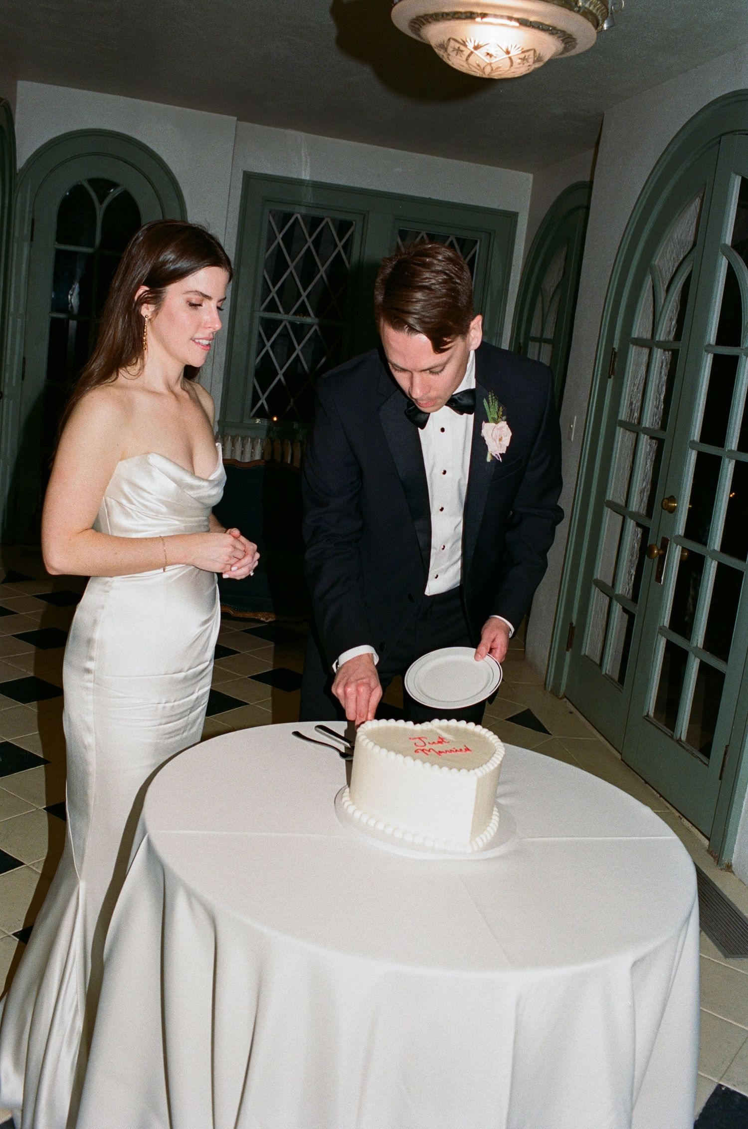 A couple cutting their white wedding cake at an elegant reception with arched doorways and chandelier lighting.