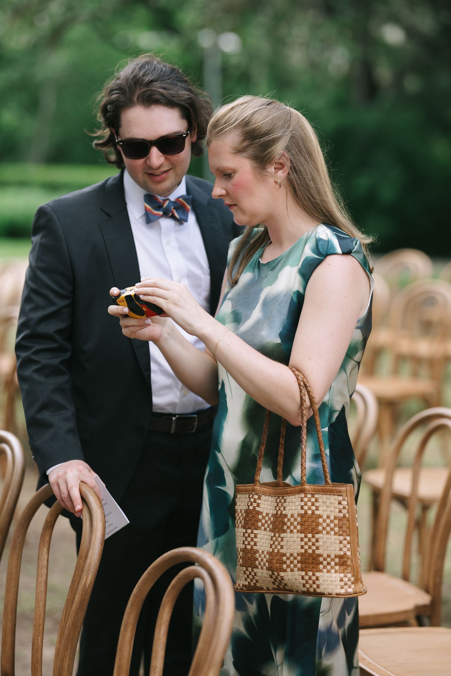 Wedding guests seated in wooden chairs examine a woven basket at an outdoor event.