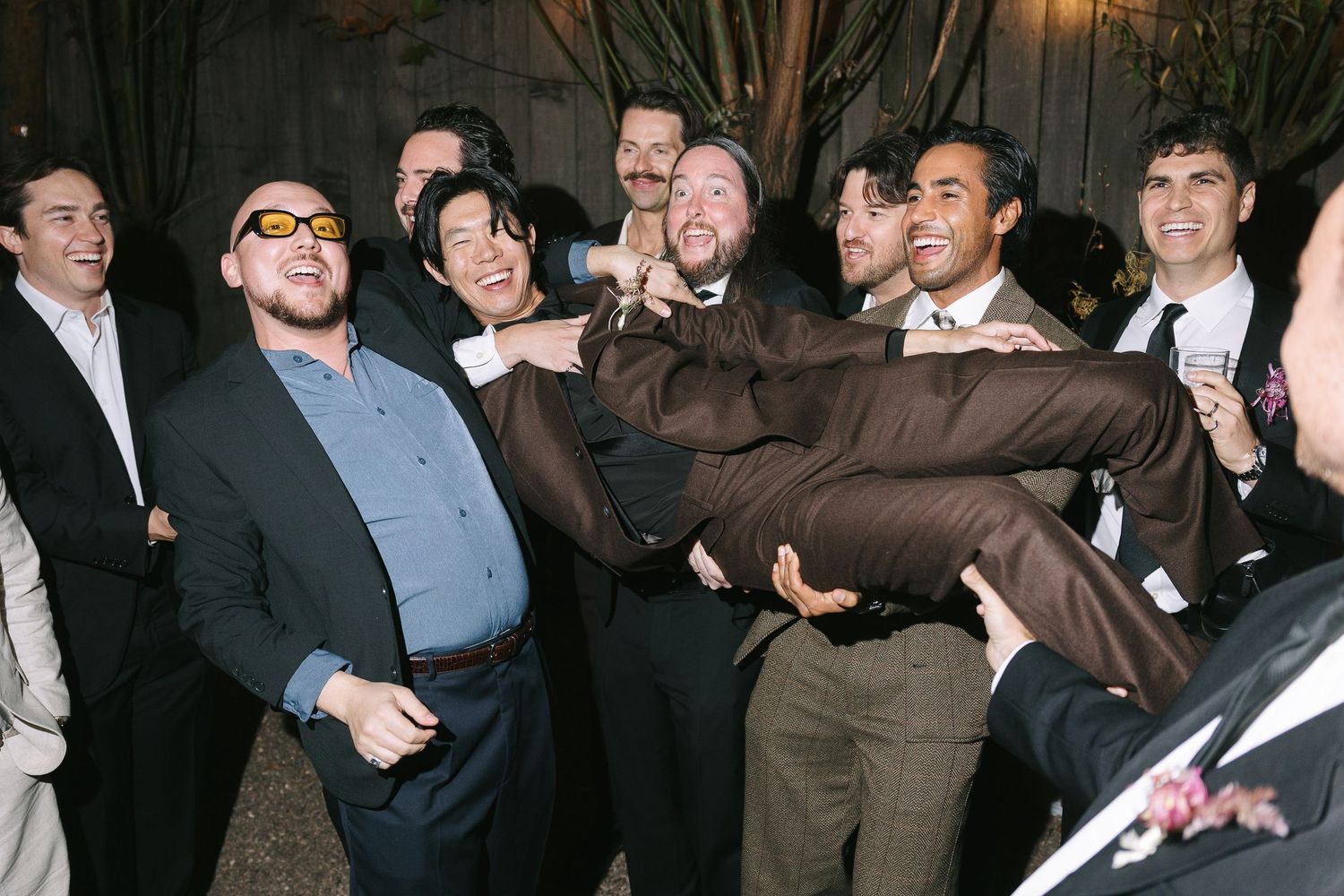 Wedding guests lift someone in celebration during an outdoor evening reception.