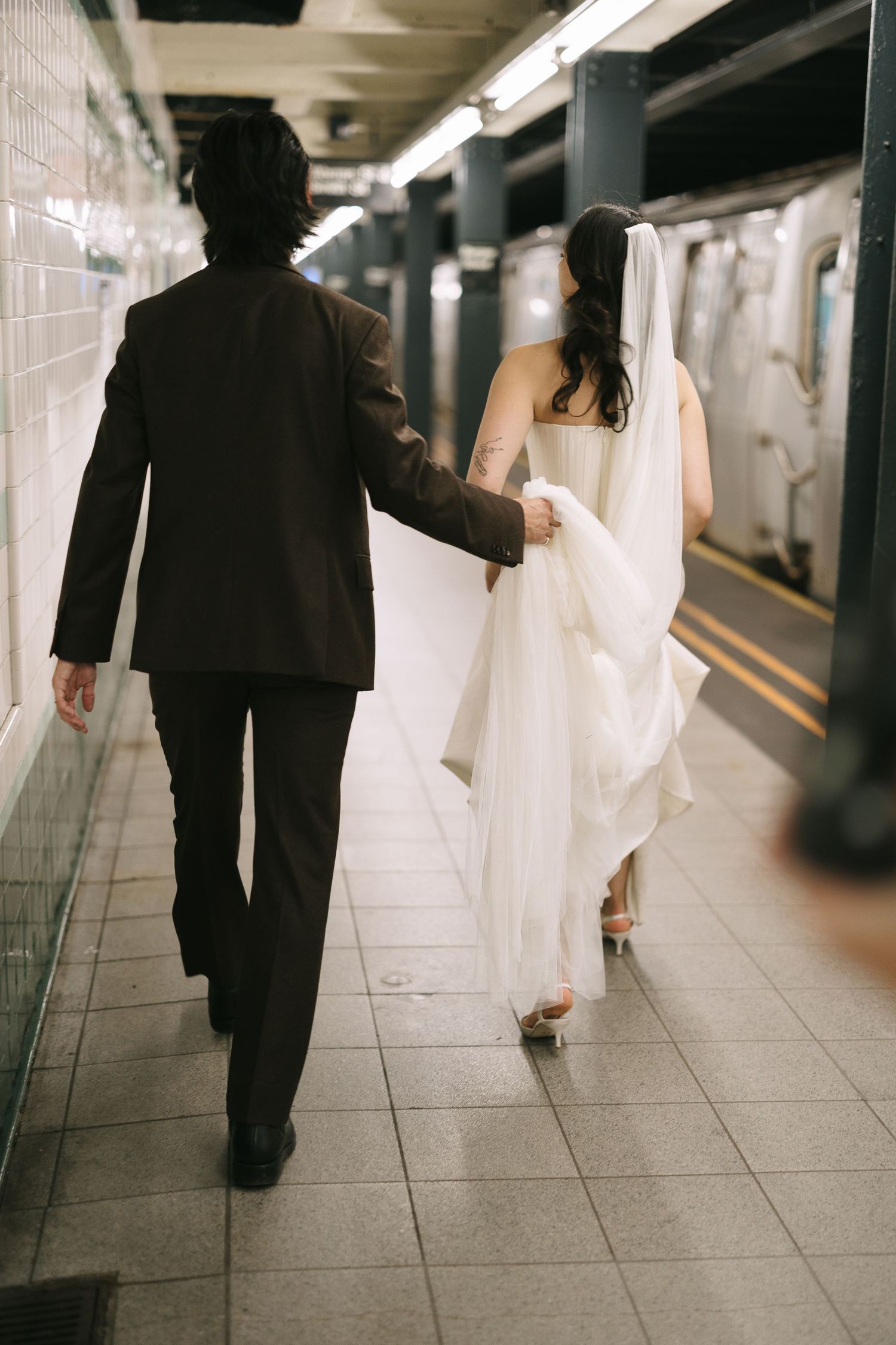Newlywed couple walking through subway tunnel, bride's dress and veil trailing behind.