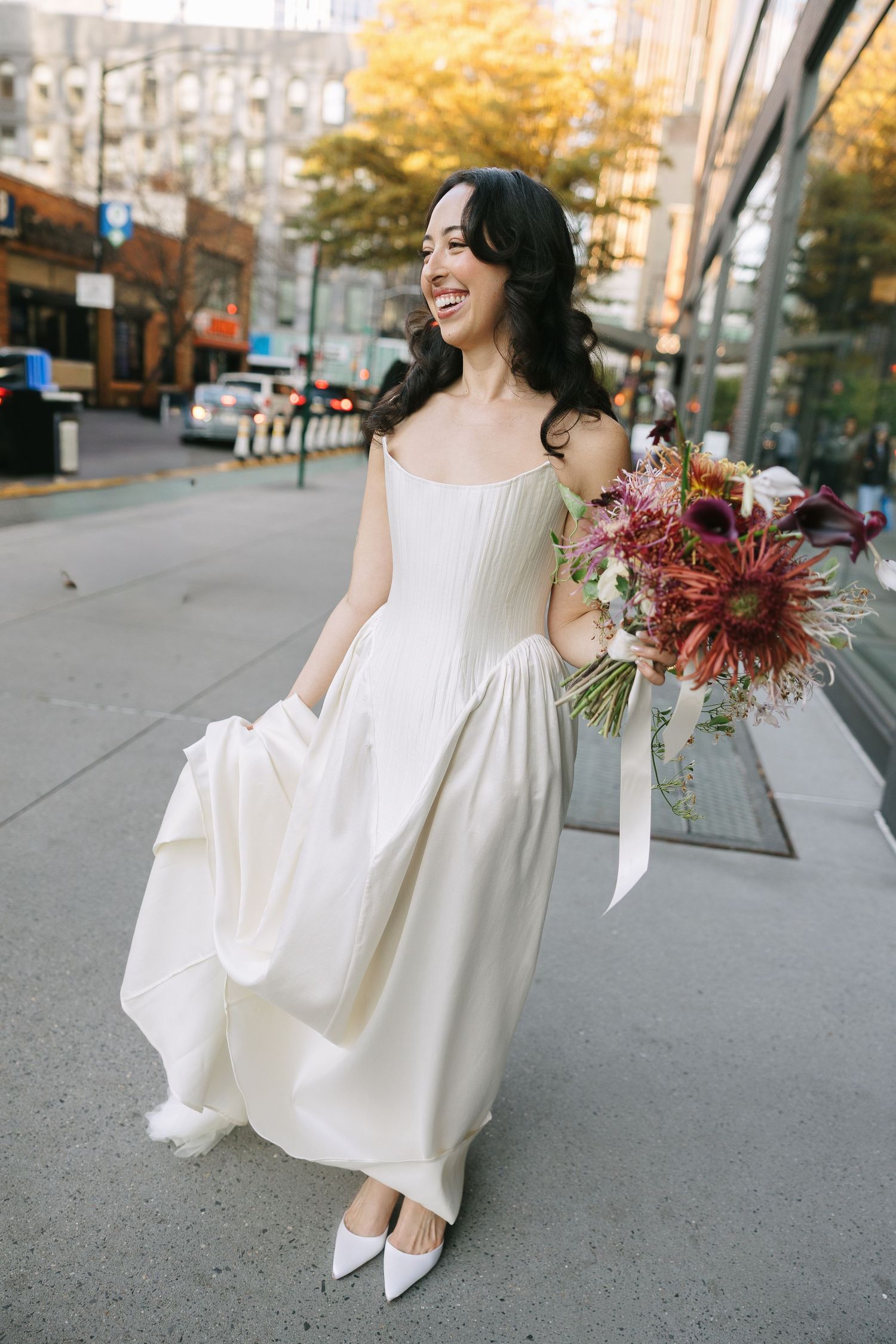 A bride in a modern white dress dances through city streets with autumn trees in background.