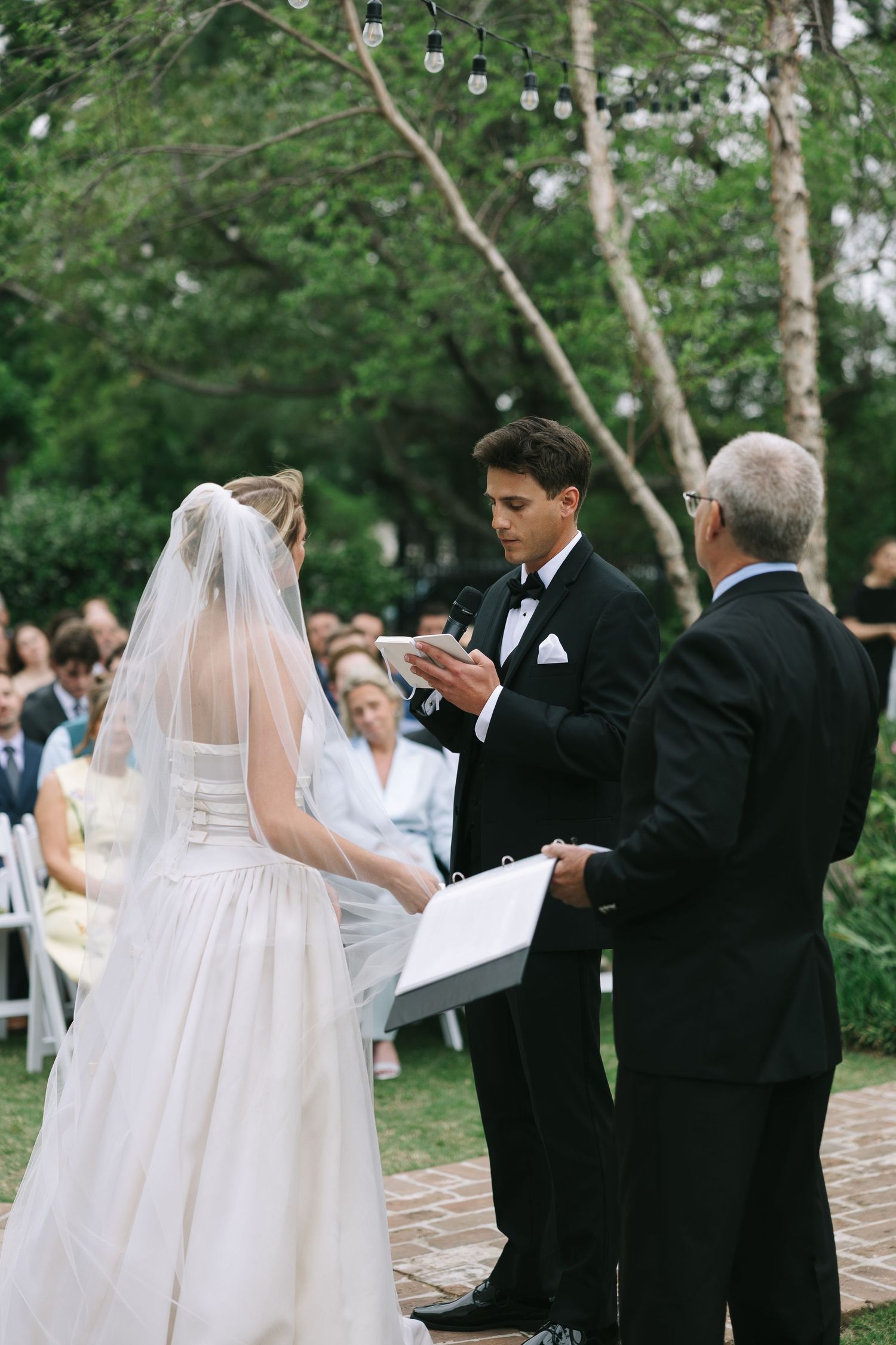 Wedding ceremony processional on a brick path with white chairs and trees in the background.