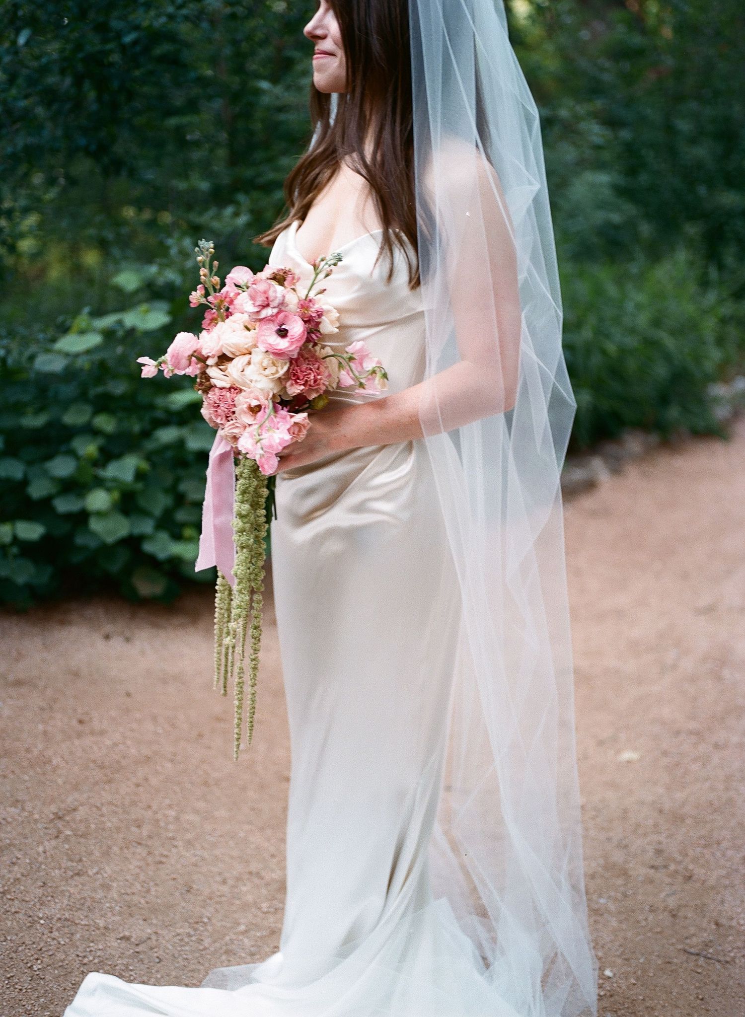 A bride in a flowing white dress holds a pink and green bouquet against a garden backdrop.