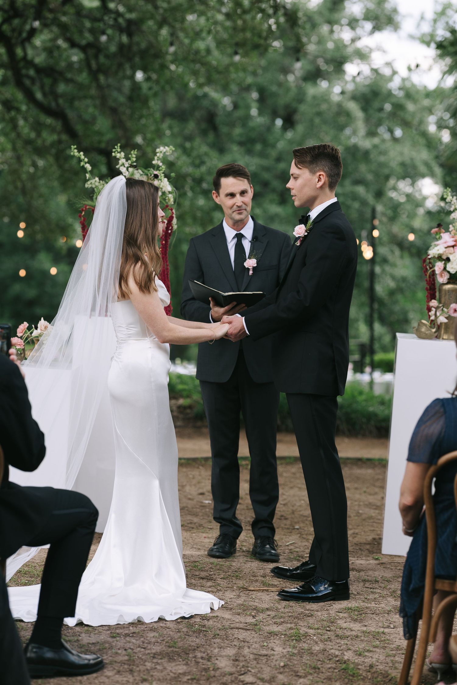 Outdoor wedding ceremony under oak trees with guests seated on wooden chairs and string lights overhead.