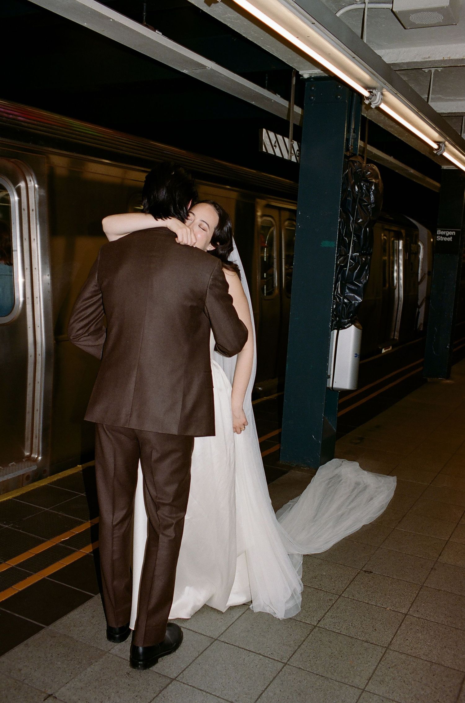 A couple in formal wedding attire walks through a dimly lit subway station platform.